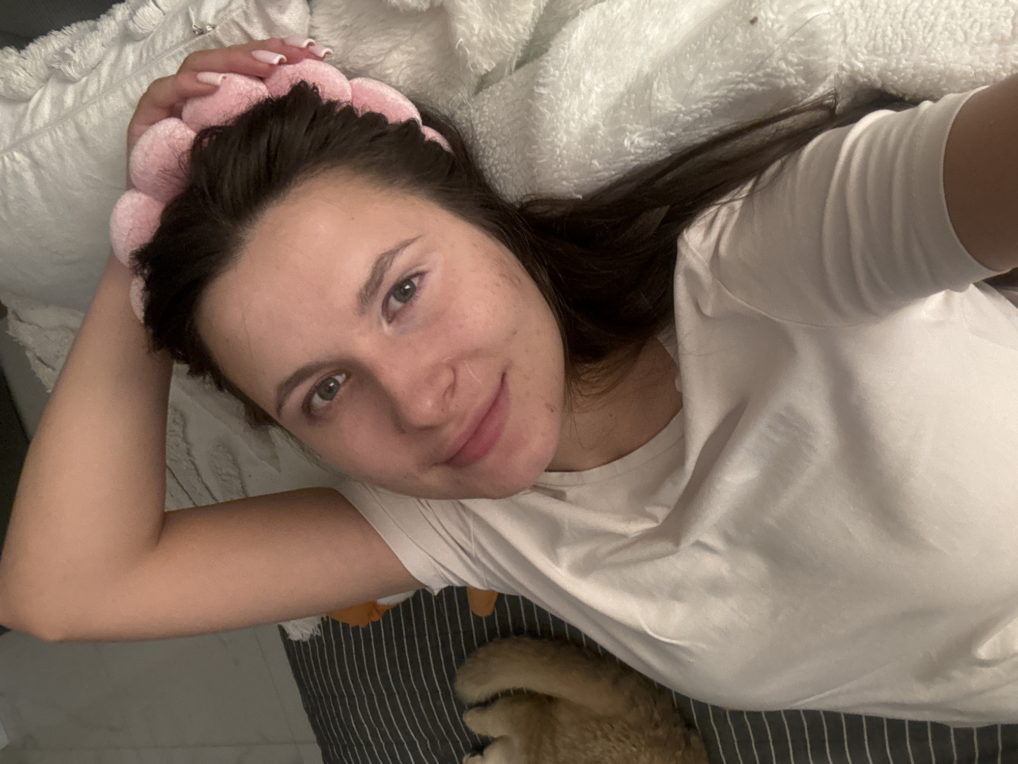 The image displays a young woman lying down in what appears to be a bedroom, capturing a selfie from an overhead perspective. She looks directly at the camera with a gentle, relaxed smile. Her dark brown hair is held back by a prominent, fluffy pink headband, potentially a skincare or comfort accessory. She is wearing a simple white t-shirt. Her right arm is bent, likely extended to hold the camera, and her nails are manicured with a light, neutral polish.
The woman has light-colored eyes and a natural complexion, with some minor skin texture or freckles visible. She is surrounded by soft bedding; a white, fluffy blanket or pillow cushions her head, and another white, textured pillow is visible near her left arm. Below her, a striped sheet or blanket, featuring dark gray or black stripes, covers the surface she is lying on. In the lower right corner of the frame, a partial view of a furry, light brown or ginger-colored pet (possibly a cat or small dog) can be seen resting on the striped bedding.
The setting suggests an indoor, private space, consistent with a bedroom. The lighting is soft and ambient, offering no specific clues about the time of day. The woman's expression conveys a sense of calm and contentment. There is no discernible text within the image, and no visual elements specifically point to the location being Odintsovo, Russia.