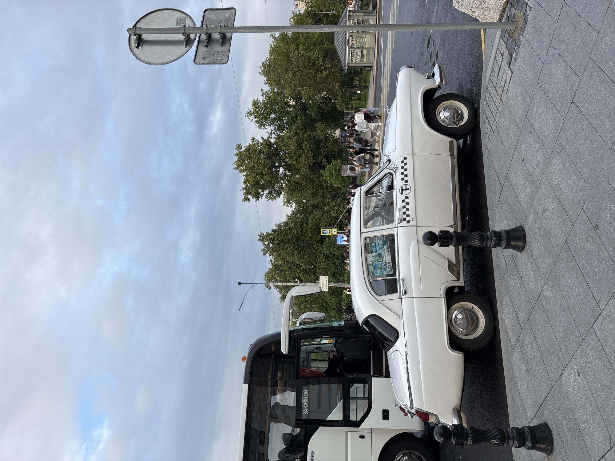 The image depicts an overcast day in a city with a mix of modern and vintage vehicles. A white, classic Soviet-era car, possibly a GAZ Volga, is parked on a street. It has a checkered taxi emblem on its side and is positioned in the foreground, partially obscuring a large, modern white tour bus. The bus has "ЗАКАЗНОЙ" (which translates to "custom order" or "chartered") written on its side in Russian.
In the background, a pedestrian crossing is visible, with several people walking across. Trees line the background, suggesting a park or a tree-lined street. A small kiosk or newsstand is also present. Traffic signs are visible, including a yellow pedestrian crossing sign. The overall impression is a street scene in a Russian city, possibly Nizhny Novgorod, given the architectural and vehicular styles. The weather appears to be cloudy and possibly humid, with damp patches on the road. The street is paved with grey stone tiles, and black bollards line the sidewalk.
