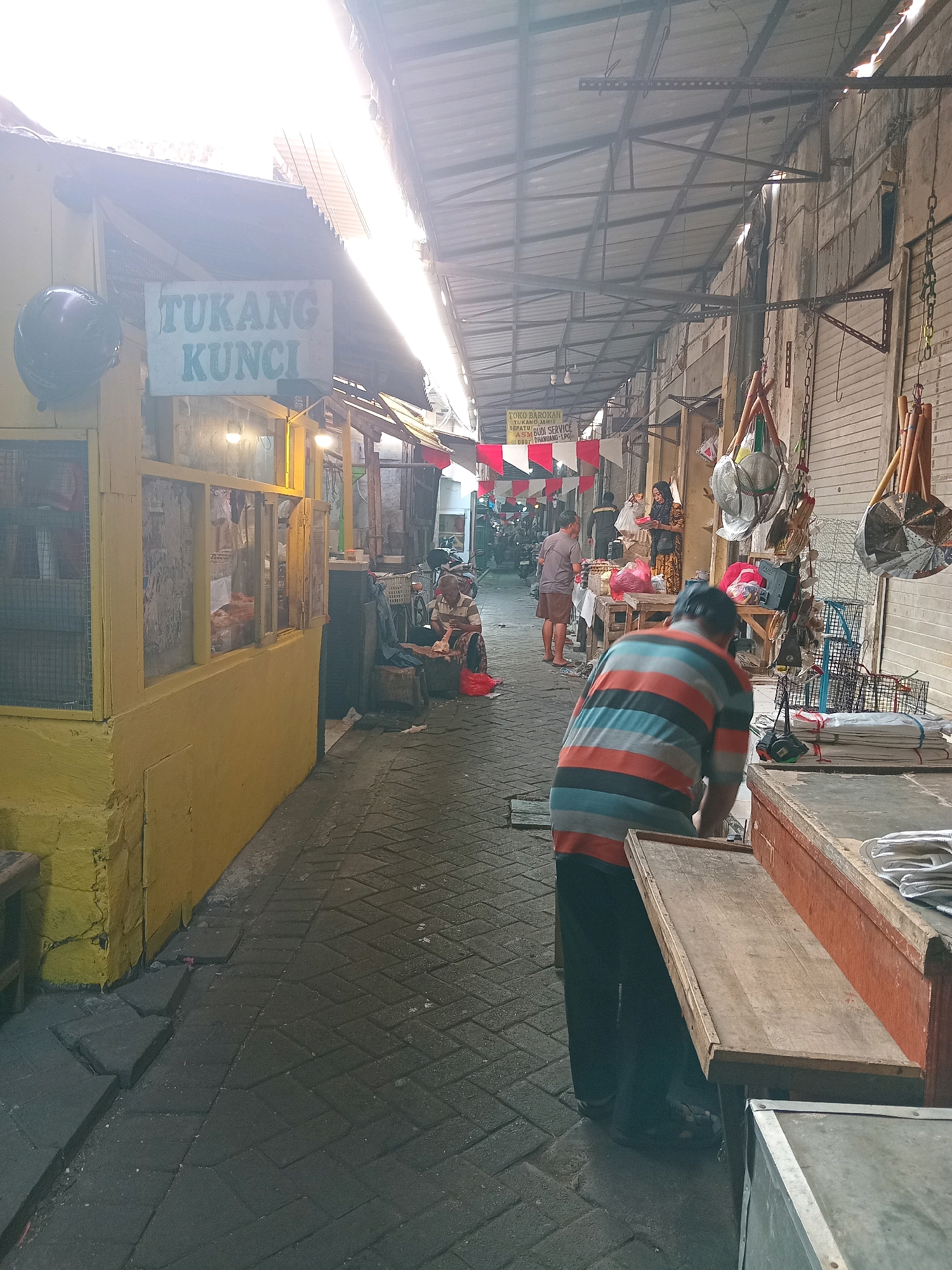 The image depicts a narrow, covered market alleyway in Pekelingan, Indonesia. The scene is dimly lit by sunlight streaming in from the right, casting long shadows. On the left, a bright yellow building with a corrugated metal roof houses a shop with the sign "TUKANG KUNCI" (Key Maker). A dark blue helmet hangs on the left side of this shop.
The alley is paved with dark grey brickwork, wet in places, suggesting recent rain or cleaning. People are present throughout the scene. In the foreground, a man wearing a striped t-shirt and dark pants is bending over a wooden counter, facing away from the camera. The counter is made of reddish-brown wood and has a wooden shelf extending from it. To the right of this counter, more wooden counters display stacked items, some wrapped in white paper.
Further down the alley, several individuals are visible. A woman in a headscarf and patterned dress is standing, holding a red bag. Another man in a light-colored shirt and shorts stands near a stall. Several market stalls line the alley, with awnings and displays of goods. Some of these goods include metal fans, baskets, and what appear to be cleaning supplies. Red and white pennants hang from a stall on the left. In the background, motorcycles are parked, indicating this is a busy thoroughfare. An elderly man sits on a stool to the right of the yellow shop, appearing to be working on something. The overall atmosphere is that of a bustling, functional outdoor market.