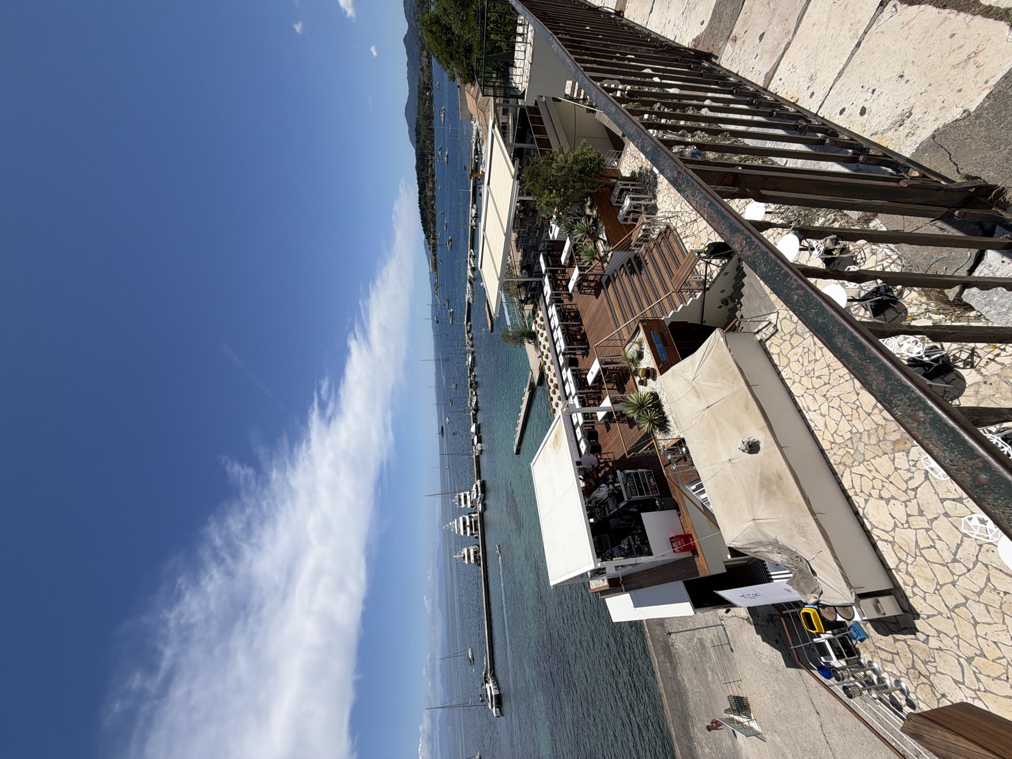 This image depicts a sunny coastal scene in Corfu, Greece, viewed from an elevated position. The clear blue sky, with sparse white wispy clouds, occupies the upper portion of the frame. Below, a tranquil expanse of turquoise water, likely a bay, is dotted with numerous boats and yachts, some moored at slender piers and others anchored further out. A verdant, mountainous coastline forms the distant horizon across the water.
In the foreground and mid-ground, a lively waterfront establishment, appearing to be a restaurant or bar, features multi-tiered wooden decks filled with tables and chairs, shaded by large white awnings and interspersed with potted plants. Several people can be observed seated and enjoying the outdoor setting. The ground surrounding the establishment is paved with light-colored, irregularly shaped stones. A robust, dark metal railing, suggesting the vantage point of the photographer, runs along the right side of the image.
The bright daylight indicates a sunny day, likely mid-morning to afternoon, with strong shadows visible across the structures. The scene conveys a relaxed, leisurely atmosphere characteristic of a Mediterranean vacation spot. A white sign with black stylized text, difficult to decipher clearly but possibly a business name or logo, is visible on one of the structures.