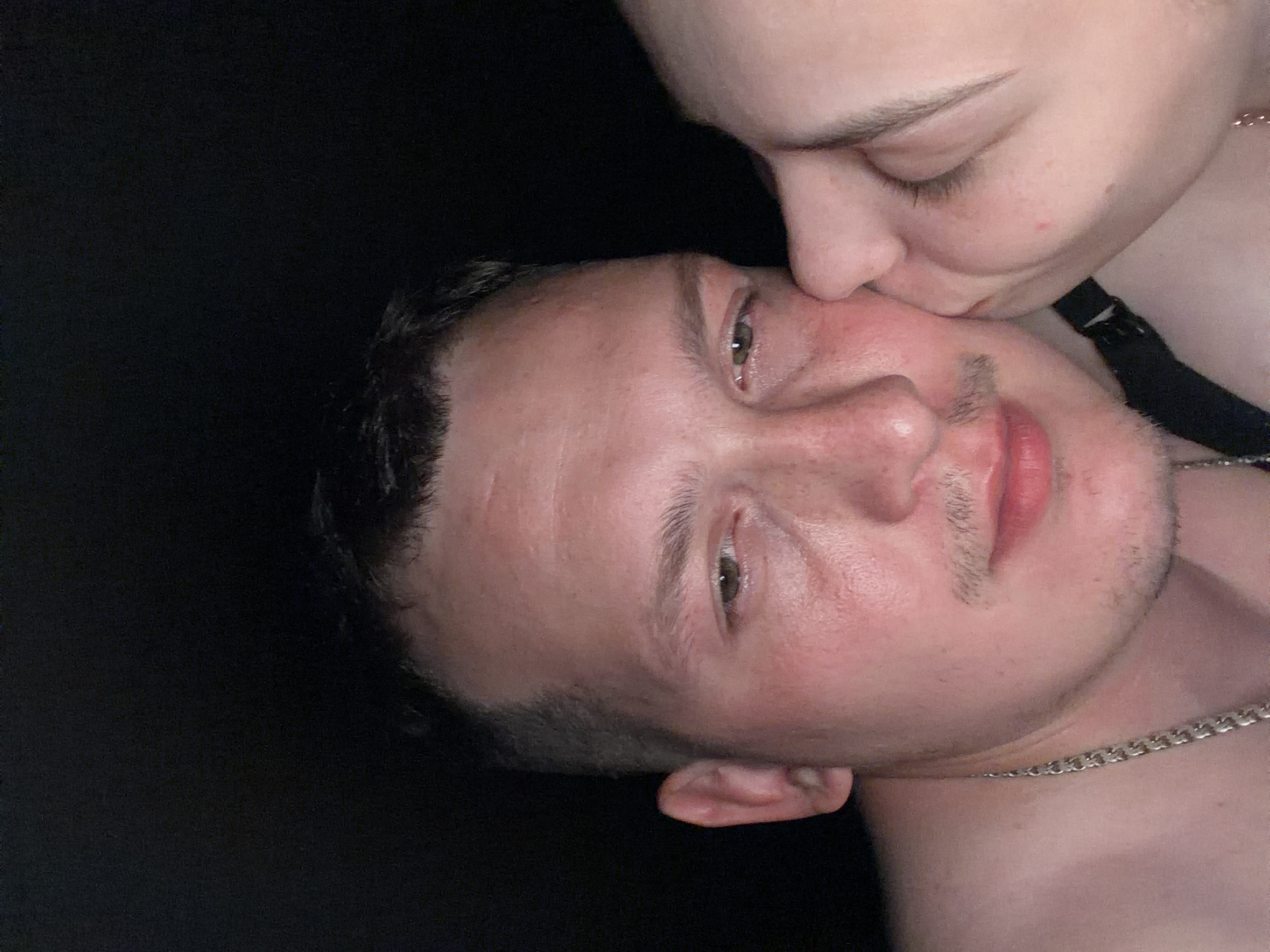The image shows a close-up of a man's face in profile, with another person's face leaning in for a kiss on his cheek. The man has short, dark hair, a light complexion, and is wearing a silver chain. The background is dark and appears to be indoors, with no discernible city or environmental features visible. There is no information within the image to suggest a location in Chaikovsky, Russia.