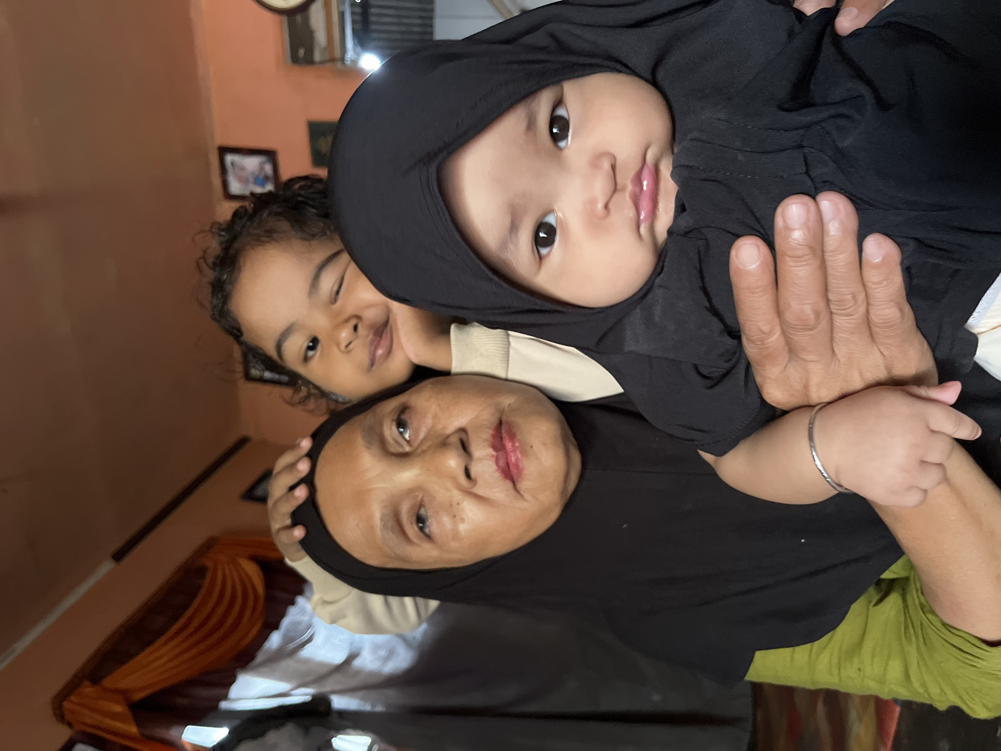 In a room with orange walls, a woman wearing a black hijab and a green shirt is holding a baby, also in a black hijab, in her arms. Behind them, a young girl with curly hair, wearing a cream-colored shirt, leans on the woman's shoulder. The room appears to be indoors, possibly a home, with curtains and framed pictures visible on the walls. The scene is likely taking place in Kruengmate, Indonesia.