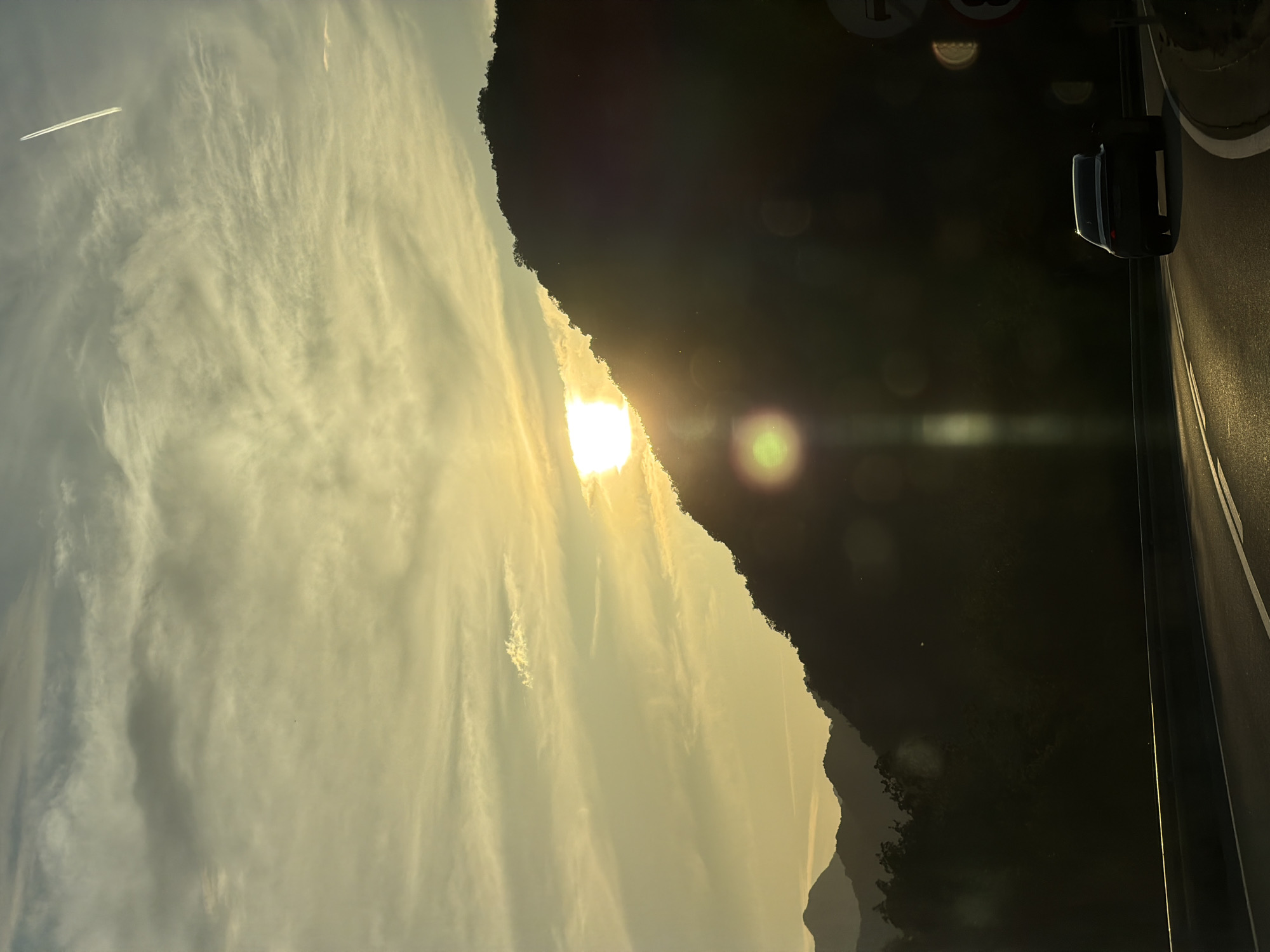 The image shows a landscape during sunset, taken from the city of Gospoština, Montenegro. The sun is setting behind a mountain range, casting a golden light onto the sky and the clouds. A contrail from an airplane is visible in the sky. On the right side of the image, a portion of a road with a street light is visible. The image appears to be taken from within a vehicle or building, as part of the frame is visible in the foreground.