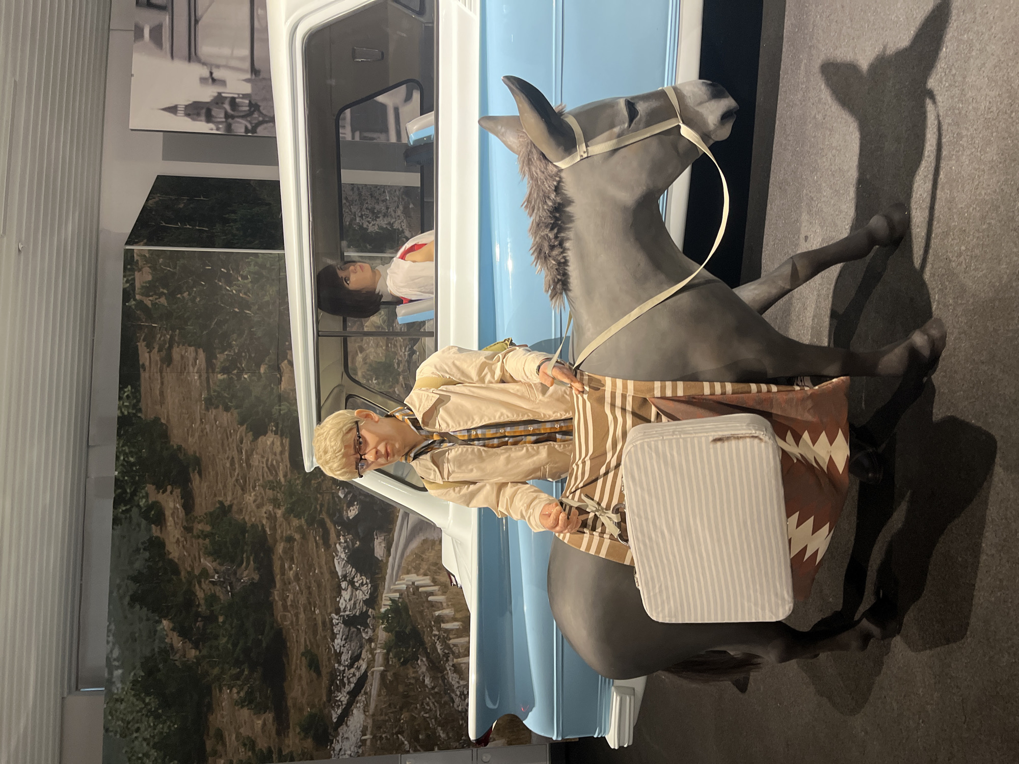 In the Museum of Military Technology of the Urals in Verkhnyaya Pyshma, Russia, a mannequin dressed in a light-colored jacket over a plaid shirt, eyeglasses, and blonde hair stands next to a mannequin donkey. The mannequin is holding a striped suitcase and a patterned blanket is draped over the donkey. Behind them, a vintage light blue and white car is visible, with another mannequin, a woman with dark hair wearing a white shirt, seated inside. The background displays a mural depicting a rugged, treed landscape with a winding road.