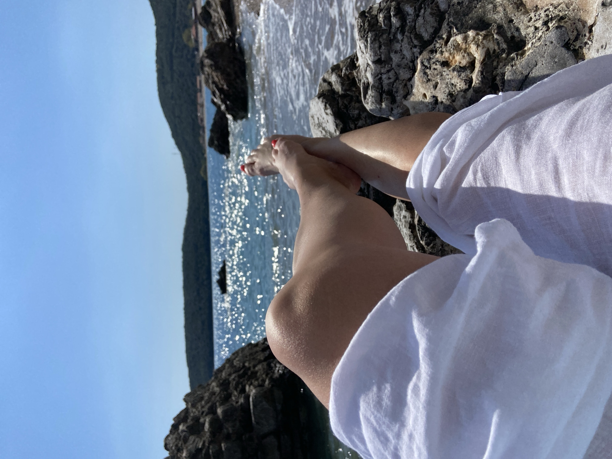 The image captures a scene on a rocky beach, likely near Prijevor, Montenegro, given the presence of similar landscapes in the region. The viewpoint is slightly elevated, looking down at a woman's legs and feet, positioned on the rocks near the water's edge. The woman's legs are bare and slightly tanned, and her toenails are painted red. She is draped in a white, semi-transparent fabric, likely a linen shirt or cover-up. The rocks are rough, dark grey, and textured with small holes and crevices. The Adriatic Sea is visible in the background, sparkling in the sunlight. Waves are lapping against the shore and rocks. In the distance, a coastline or another rocky formation is seen, covered with vegetation. The sky is a clear blue. The overall atmosphere is bright and sunny, indicative of a summer day.
