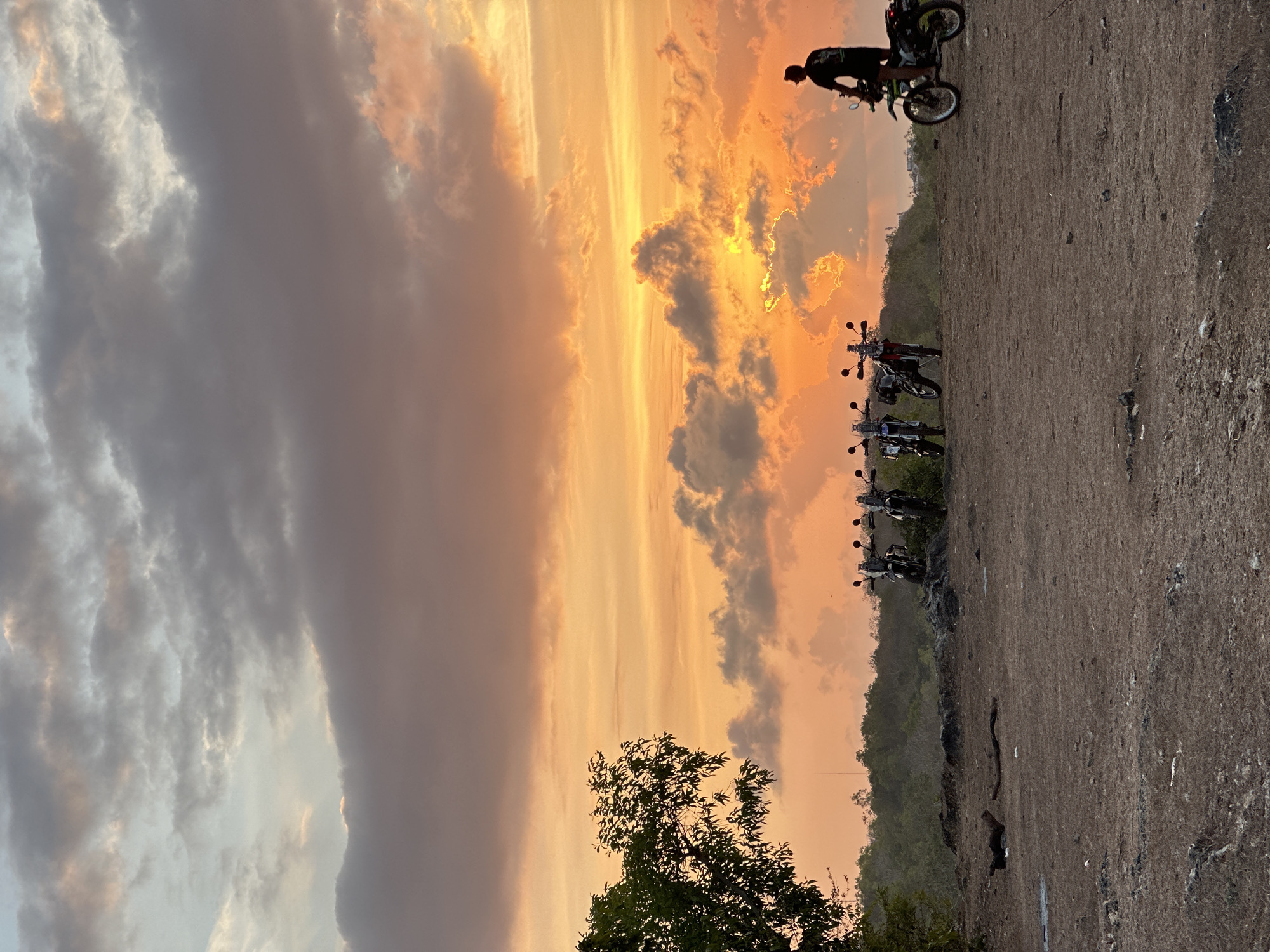 A vertical, full-shot image captures a dramatic sunset over a coastal landscape in Kuta Selatan, Indonesia. The sky is filled with vibrant hues of orange, yellow, and pink, with dark gray clouds creating a stark contrast. The sun is partially obscured by clouds, casting a warm glow across the scene.
In the foreground, a dirt and gravel area is sparsely covered with dry grass and a few pieces of debris, including sticks and rocks. To the left, a dense green tree stands tall.
Further back, a line of four motorcycles is parked on a slightly elevated rocky outcrop, facing left. A person wearing a dark shirt and shorts is seated on the rightmost motorcycle, which is pointed towards the right and appears to be in motion, with its rear wheel kicking up dust. Another person is partially visible in the distance, standing near the motorcycles.
Beyond the motorcycles, a dense green treeline stretches across the horizon, with subtle hints of buildings and structures visible in the far distance. The overall impression is one of a rugged, natural landscape bathed in the warm light of a setting sun.