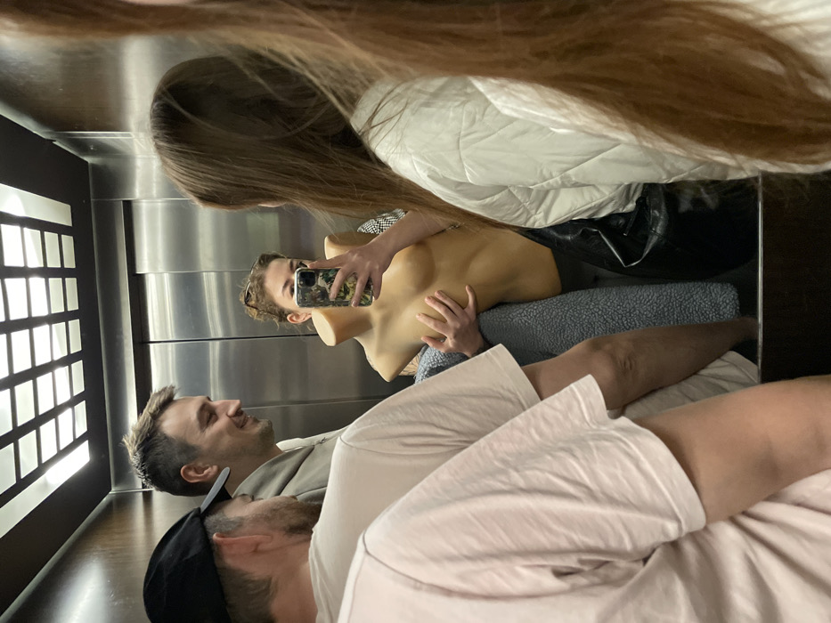 In an elevator selfie, a woman holds a mannequin torso. The mannequin is nude from the shoulders up. The woman's reflection shows her with long brown hair. Other people in the elevator are visible: one man is looking up at the woman, and another man with a black baseball cap is looking down. The elevator has a mirrored wall, and windowed doors. The light in the elevator is bright.