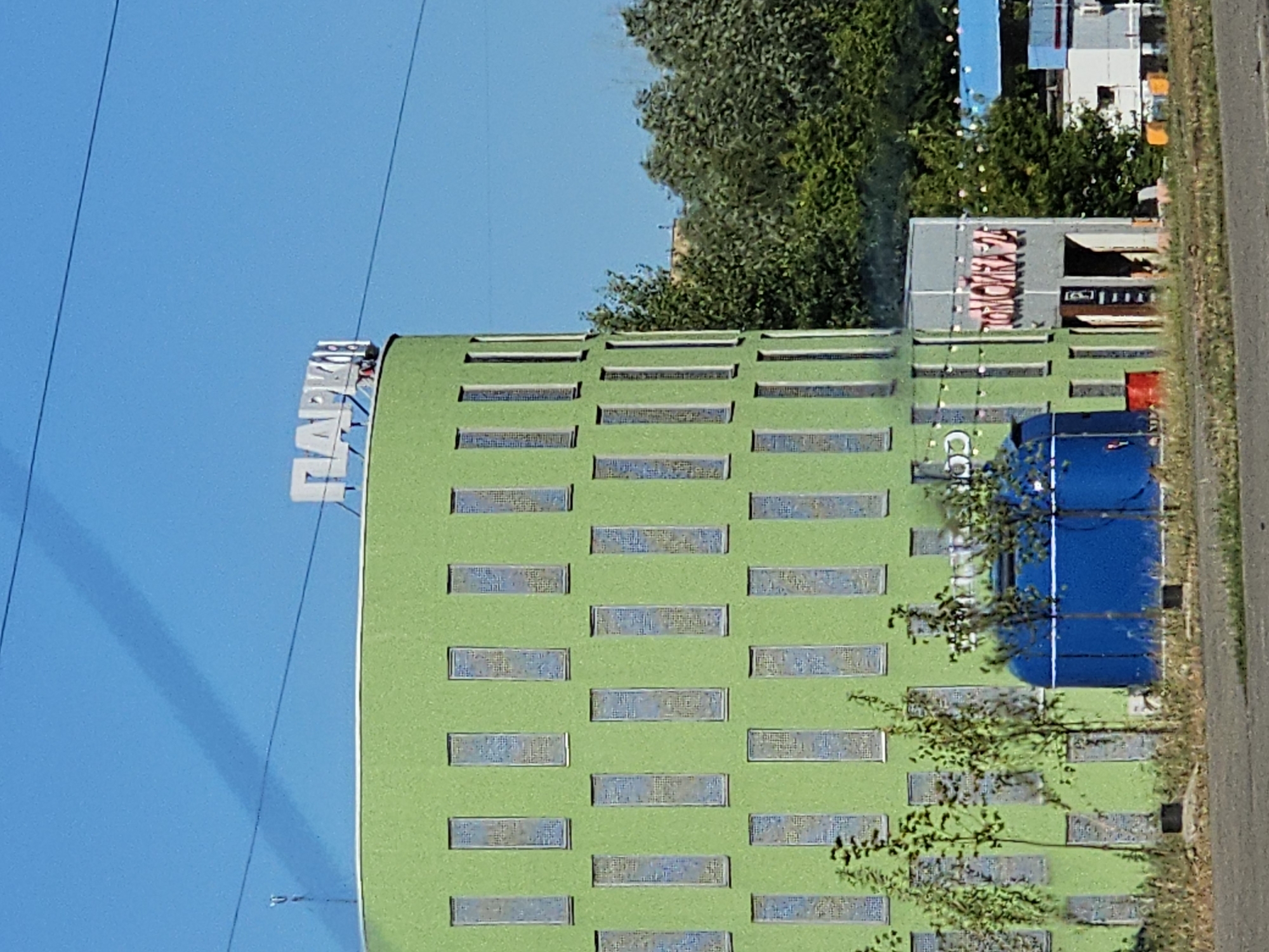 The image shows a modern, lime-green building with rows of rectangular windows. The building is oriented vertically in the frame, suggesting it's a tall structure. On the left side of the building, a large white sign with blue letters spells out "TAXI". In the background, a large tree with dense green foliage partially obscures another building that appears to be a light grey structure with a sign that has pink lettering. To the right of the green building, a bright blue rectangular container or structure is visible. The sky is a clear, bright blue, indicating it is daytime. A shadow from overhead wires falls diagonally across the blue sky on the left side of the frame. The scene appears to be an urban or commercial area, possibly a hotel or office complex, given the modern architecture and signage. The overall impression is one of a sunny day in a contemporary setting. The image is a photograph. No people are visible in the frame.