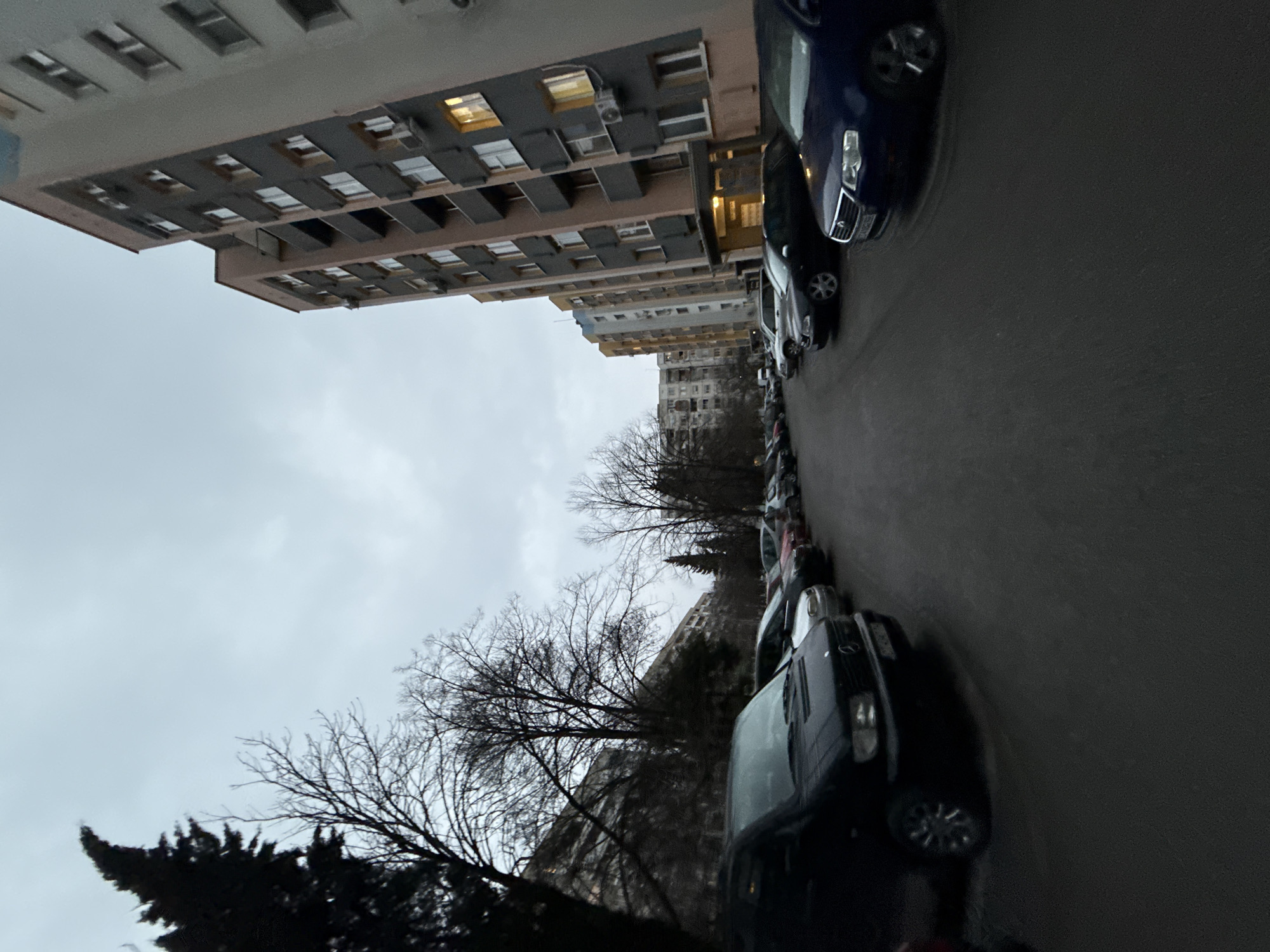 The image depicts a street scene in Stara Zagora, Bulgaria. The view is along an asphalt road, where cars are parked in a row on the right side of the frame. Buildings of various heights are on the left and in the background. The sky is overcast. Trees with bare branches are visible along the roadside.