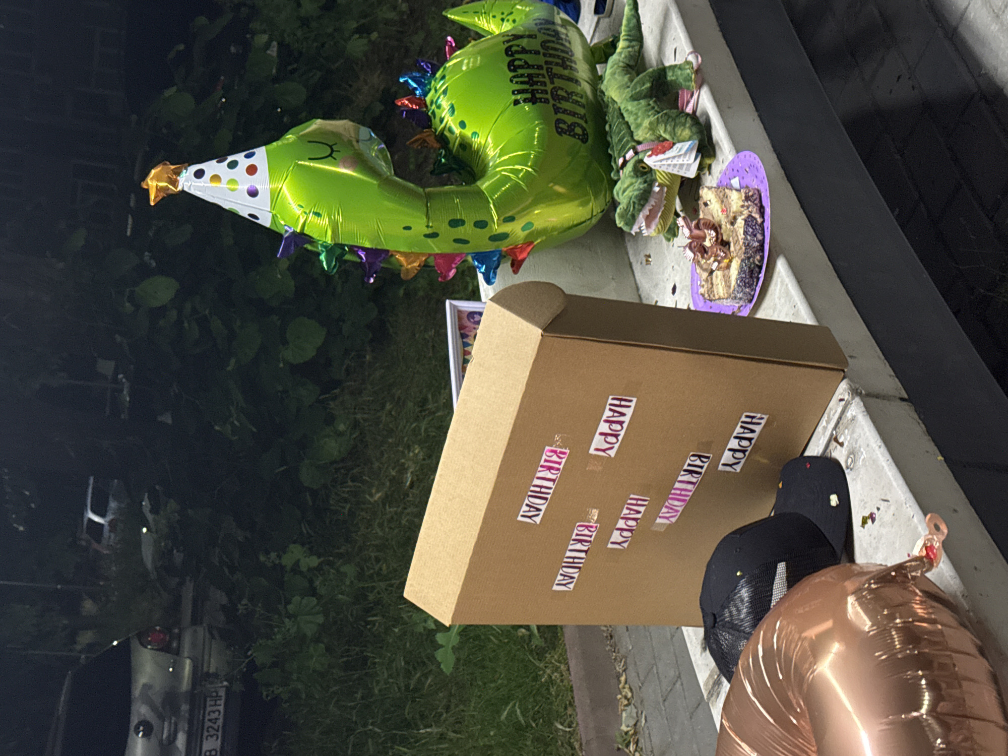 A still life scene is set outdoors at night, illuminated by artificial light. In the foreground, a large, rose-gold balloon lies on its side, partially obscuring a black baseball cap. To the right, a cardboard box is propped open, its exterior decorated with pink and white stickers that spell out "HAPPY BIRTHDAY" multiple times. Next to the box, a bright green dinosaur-shaped balloon, wearing a party hat, stands tall. Below the dinosaur balloon, a slice of cake with a golden "5" candle sits on a purple plate. A green plush alligator toy is positioned behind the cake. In the background, several vehicles are visible, including a silver car with a license plate reading "B 3243 HP," parked near lush green foliage and a paved walkway. The overall scene suggests a birthday celebration.