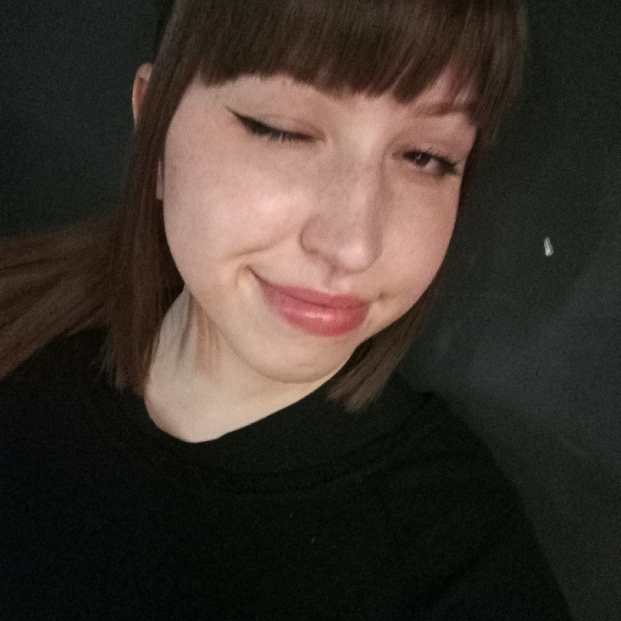 A young woman with fair skin, brown hair, and bangs is smiling and winking at the camera. She has eyeliner on her upper eyelid. She is wearing a black shirt. The background is a dark, textured surface. There is no information available regarding the location as Chaikovsky, Russia.