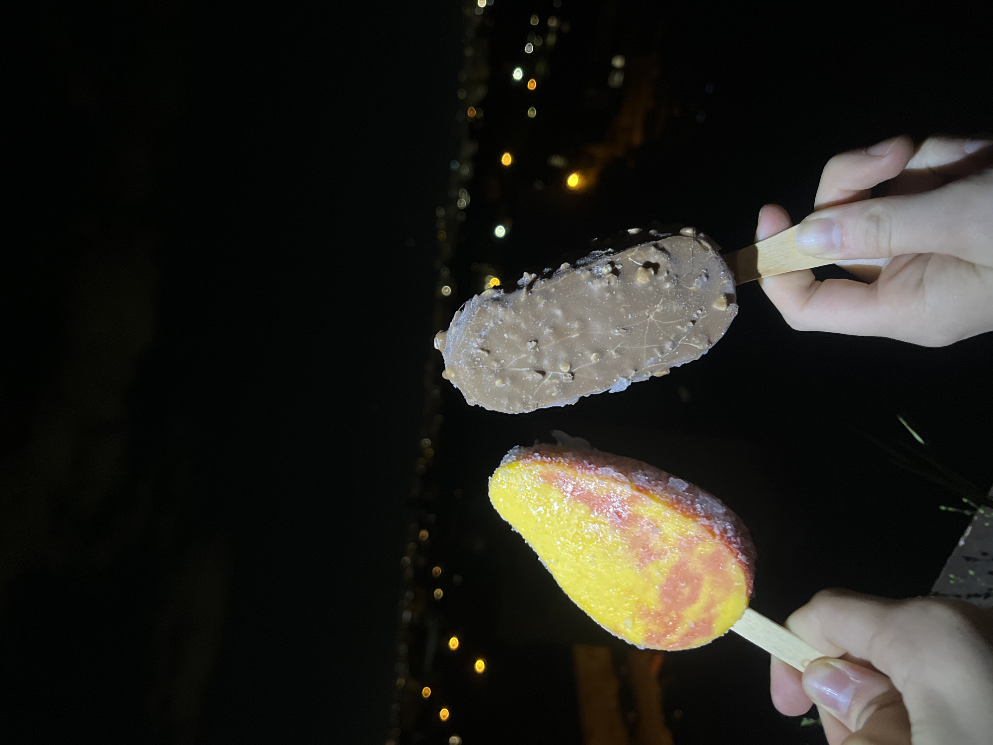 Two hands hold ice cream popsicles on sticks at night. The upper popsicle is brown with nuts. The lower popsicle is yellow and pink. In the background is a blurry cityscape with lights. The location is Sofia, Bulgaria.
