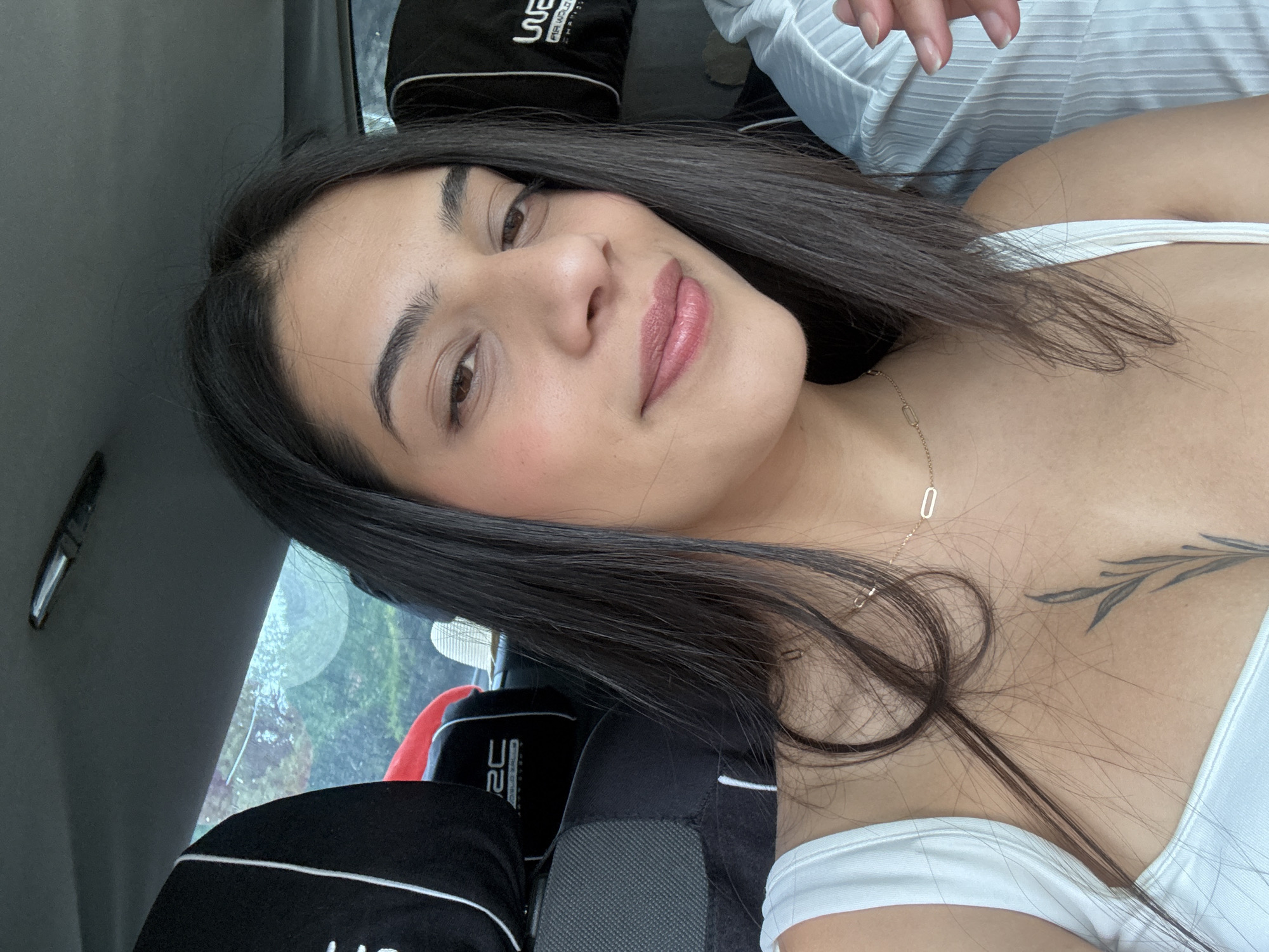 A woman with dark hair and light skin is positioned in the front seat of a car. She is wearing a white top. A silver necklace with a rectangular pendant is visible around her neck. A black and white car seat cover with the letters "SC" is partially visible. Through the car window, green foliage and blurry shapes suggest an outdoor environment. There is a tattoo of leaves on her chest. The city of Unknown, Unknown is not visible.