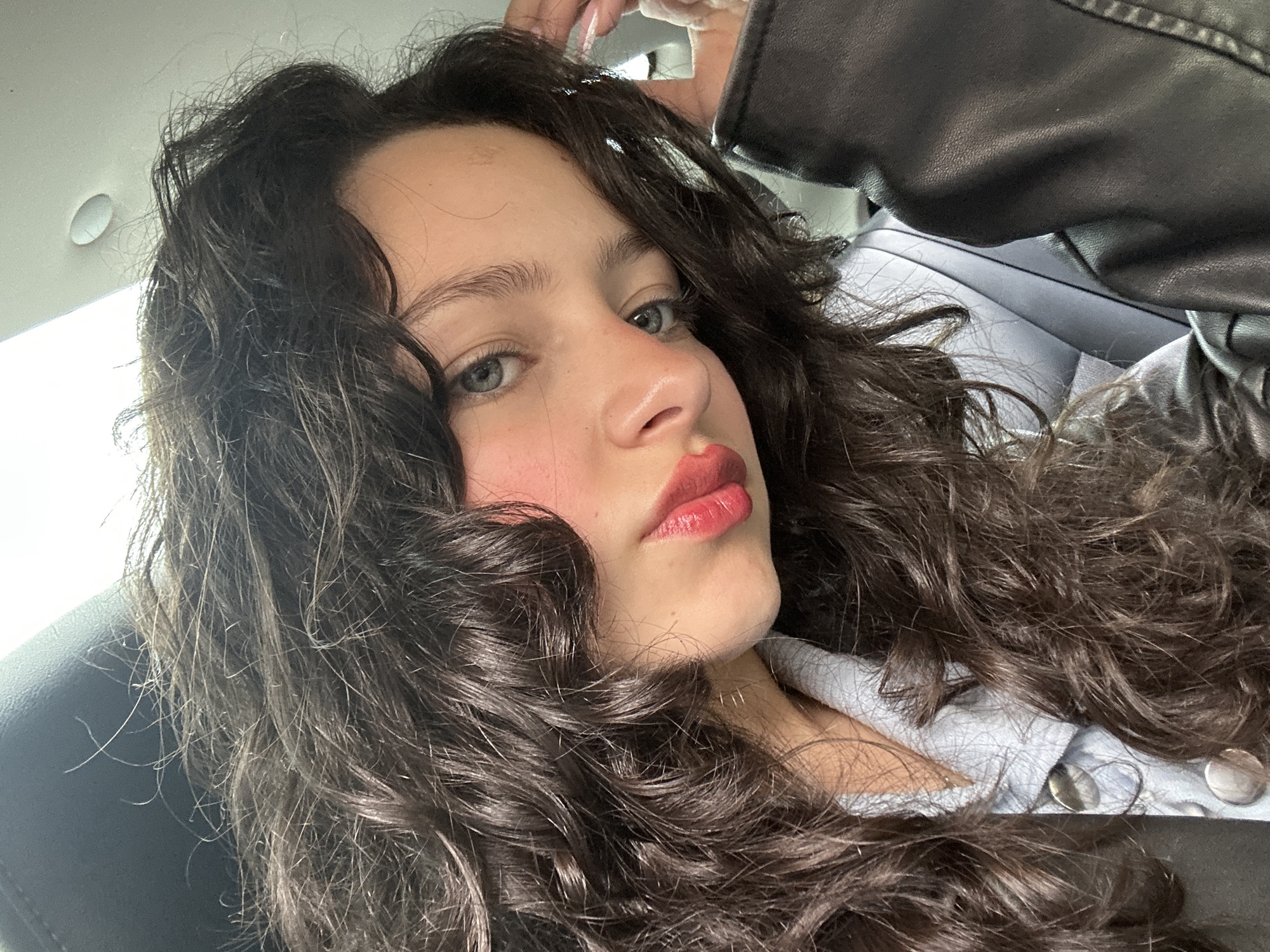 A young woman with curly, dark brown hair and fair skin is shown in a close-up selfie. She has blue eyes and is wearing red lipstick and what appears to be blush. She is looking at the camera with a slight frown. Her left arm is raised, with her hand near her head. She is wearing a black leather jacket. The background shows part of a car interior, including a gray car seat and part of the car ceiling. The setting appears to be inside a car. There is no clear evidence to confirm the location is Yerevan, Armenia.