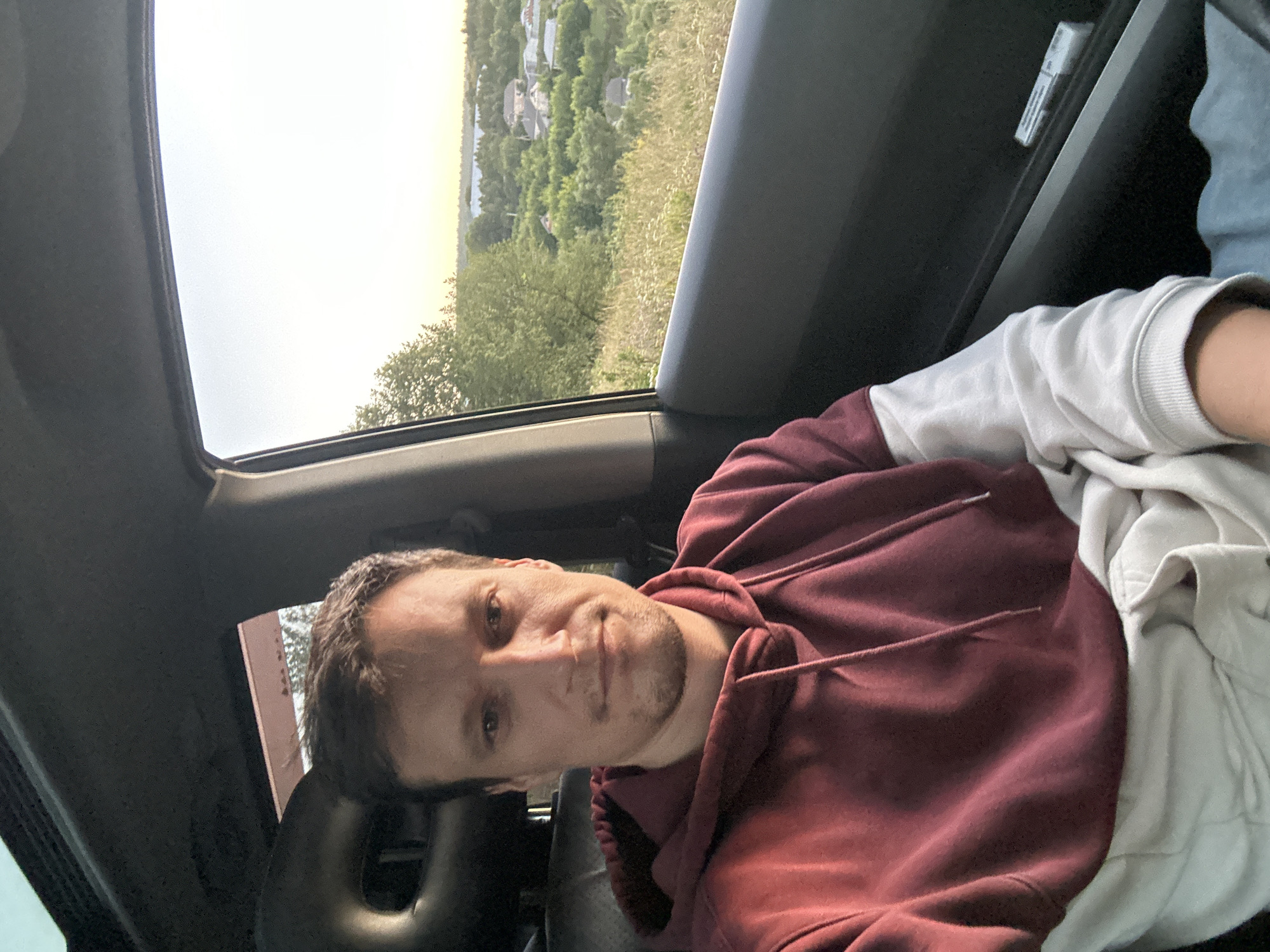 A selfie is taken from inside a car by a man wearing a maroon and white hoodie. He is looking directly at the camera with a neutral expression. The car interior is visible, including the headrest and a portion of the dashboard. Outside the car window, a scenic view of a rural landscape with trees, houses, and a body of water can be seen. The setting appears to be on the outskirts of Makiv, Ukraine, during daylight hours.