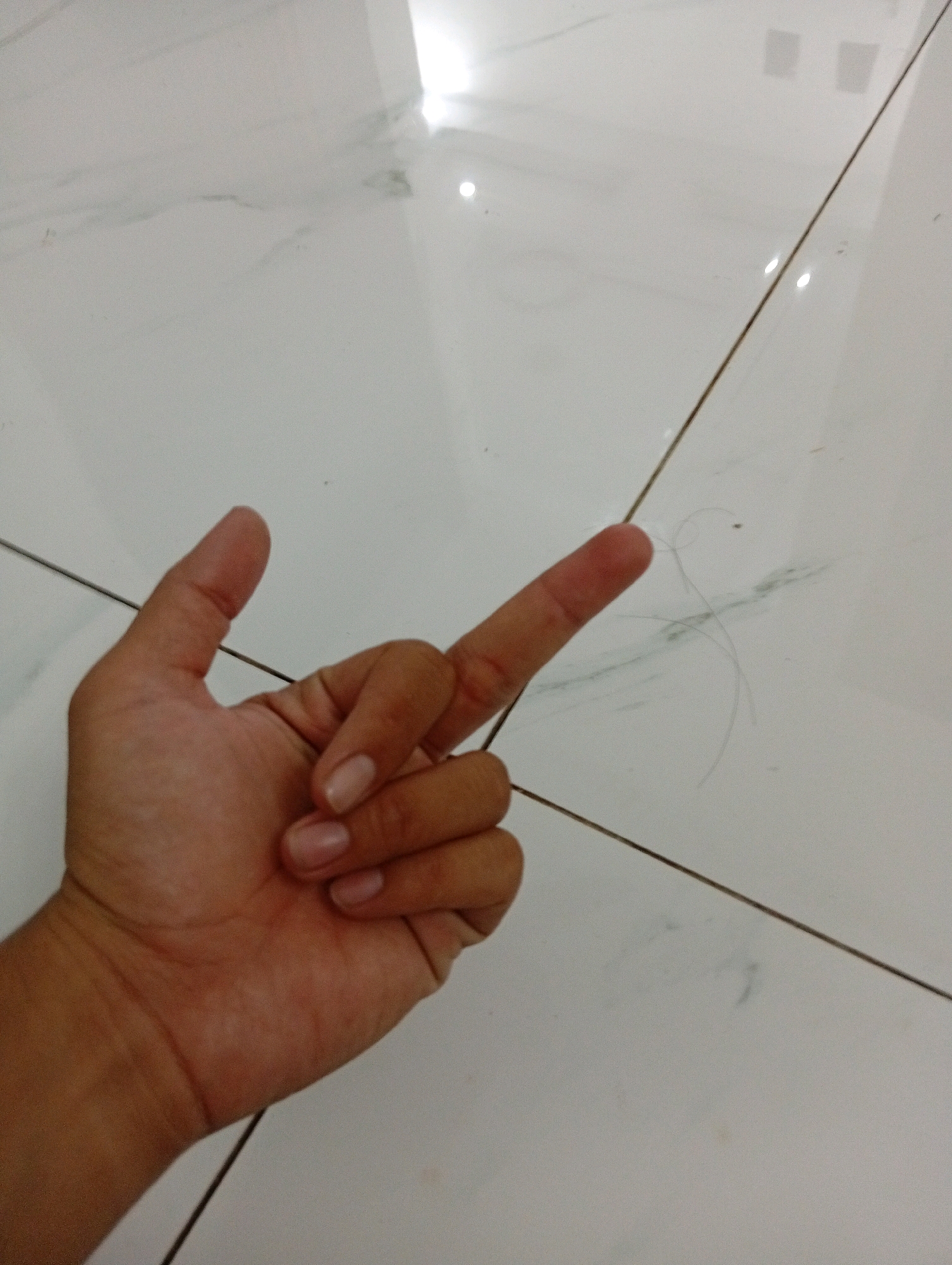 This media file features a close-up view of a person's hand, with the palm facing upwards and fingers positioned to form a "rock on" gesture. The hand is positioned on a tiled floor. The tiles appear to be white with subtle grey veining, creating a slightly reflective surface that shows a few small, light reflections, possibly from artificial lighting. A thin dark line, likely grout or a seam between tiles, runs diagonally across the image. A few strands of light-colored hair are visible on the floor near the fingers, suggesting a domestic or indoor setting. The lighting appears to be artificial and somewhat bright, as indicated by the reflections on the tiles. There is no visible text or explicit indication of the time of day or weather. The overall impression is a casual, everyday moment captured from a low angle.