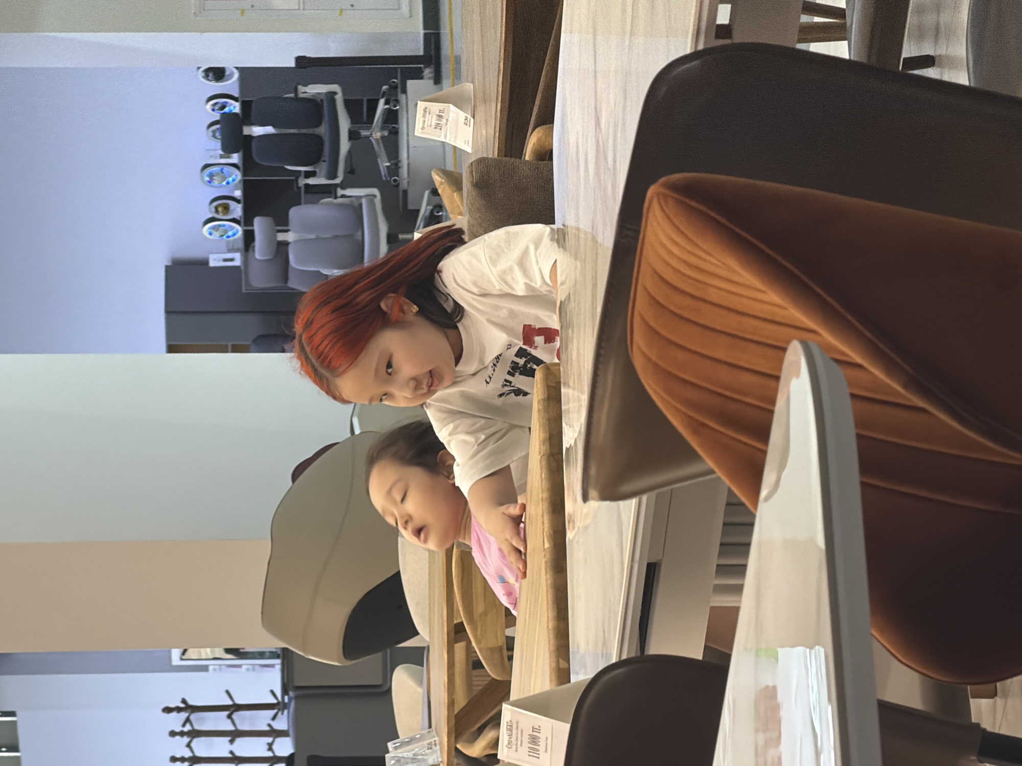 The image depicts two young girls in what appears to be a furniture store. The girl in the foreground, on the right, has bright red hair and is wearing a white t-shirt with red lettering. She is looking to her left and smiling. To her left, the second girl is lying down with her eyes closed, wearing a pink shirt. They are seated at a wooden table with a marble-like surface. In the background, various furniture items are visible, including chairs and tables, and some price tags. The environment suggests a retail setting, possibly in Astana, Kazakhstan, given the presence of Cyrillic script on some of the price tags which are common in the region. The lighting is bright, indicating an indoor commercial space.