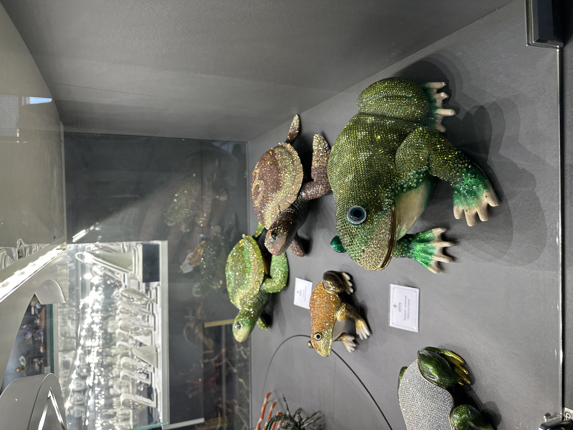 The image shows a display case in Saint Petersburg, Russia, featuring jeweled figurines of amphibians and reptiles. There are two turtles and three frogs. One frog is large and green with a prominent blue eye, another is smaller and golden, and the third is smaller and green with a silver top. A black wire runs across the bottom of the display. There are price tags under the frogs. The inside of the display case contains jewelry.