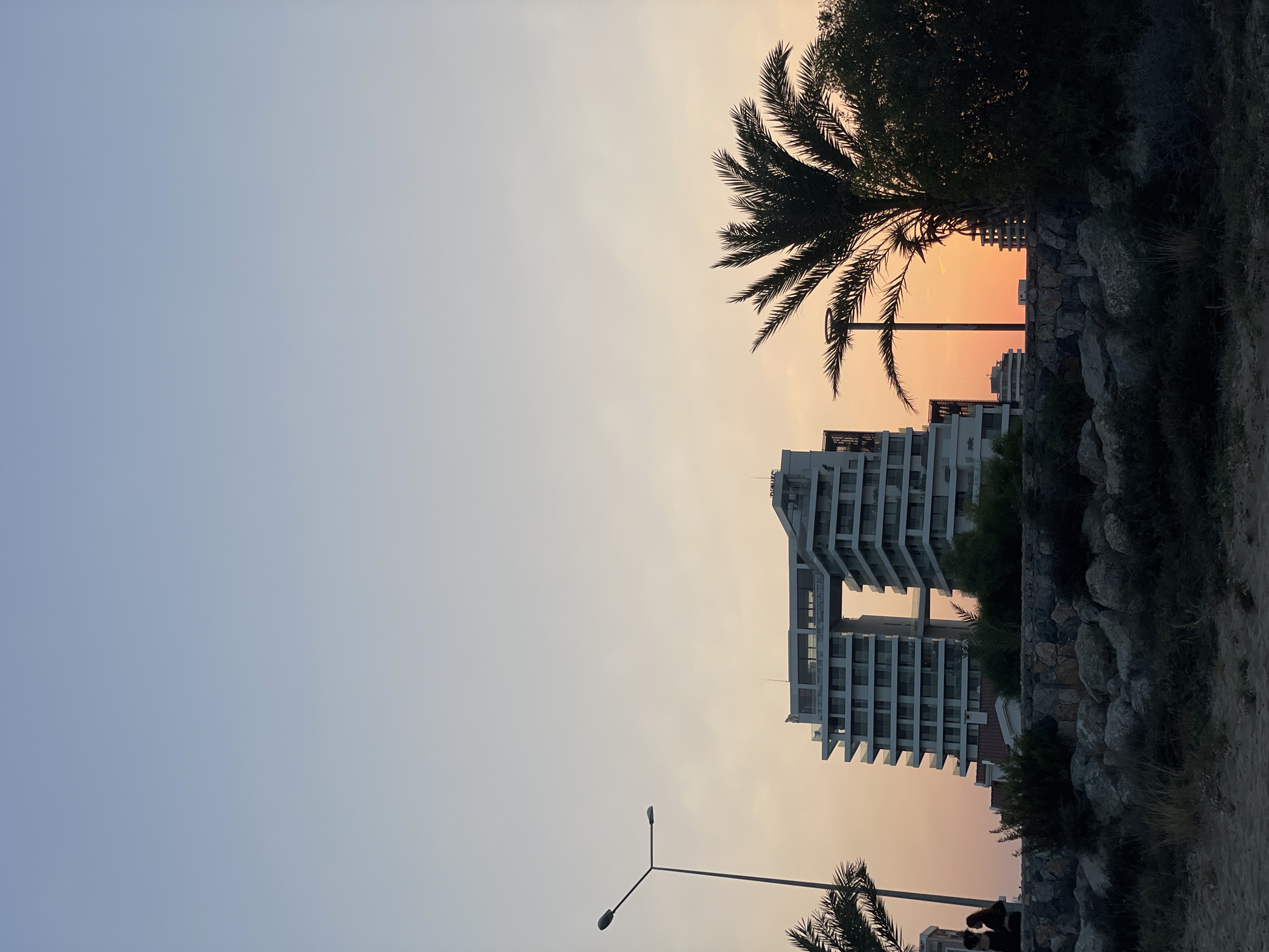 A wide shot captures the sunset over a coastal area in Trikomo, Northern Cyprus. The sky transitions from a pale blue at the top to warm orange hues near the horizon. A modern, angular white building with a large central opening stands prominently in the middle ground, its geometric design silhouetted against the sky. To the left of this building, a tall, slender street lamp with two lights is visible. A silhouetted palm tree with its fronds spread wide is positioned to the right of the main building, obscuring part of the sunset. In the foreground, a rocky embankment covered in dry vegetation separates the beach from the buildings. A small group of people, two of whom are standing close together, are visible on the left side of the embankment, near a palm tree. The beach in the immediate foreground is made of sand and scattered dry grasses.