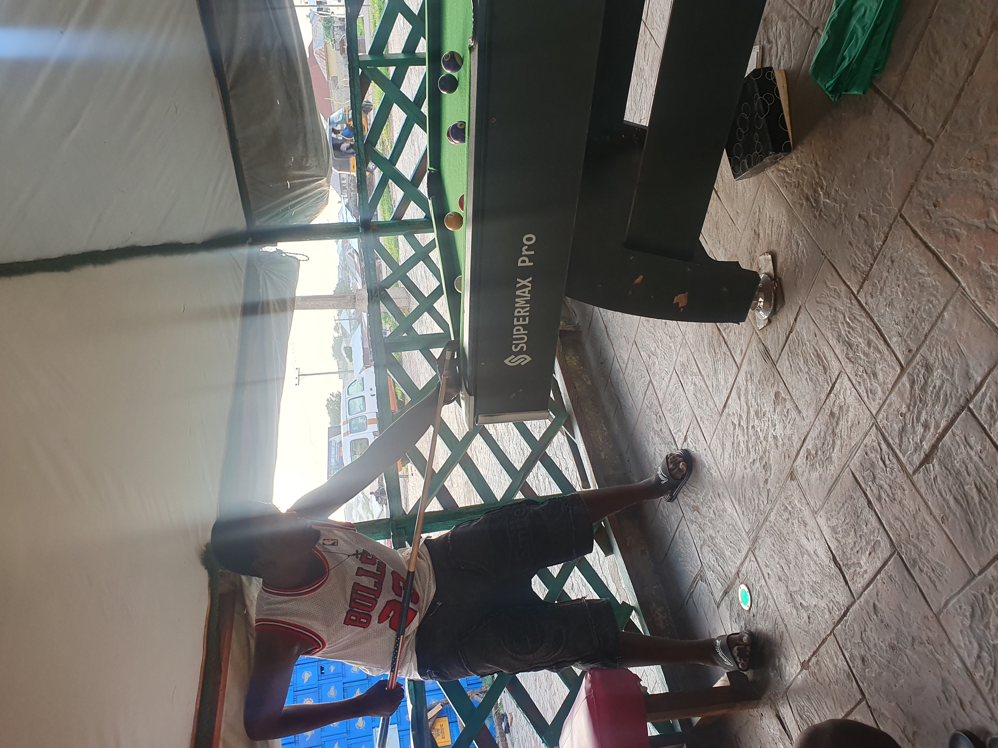 A young man wearing a white Chicago Bulls basketball jersey with the number 23 and dark shorts is playing pool. He is holding a pool cue and aiming at a ball on a dark green pool table labeled "SUPERMAX Pro". The pool table has several balls on it, including yellow, purple, and black balls. The scene appears to be outdoors under a canopy, with a patterned tiled floor. In the background, there is a green wooden fence, some buildings, and vehicles on a street, suggesting an urban environment. The lighting suggests it is daytime, possibly late afternoon. The presence of a red stool next to the pool table indicates a seating area.