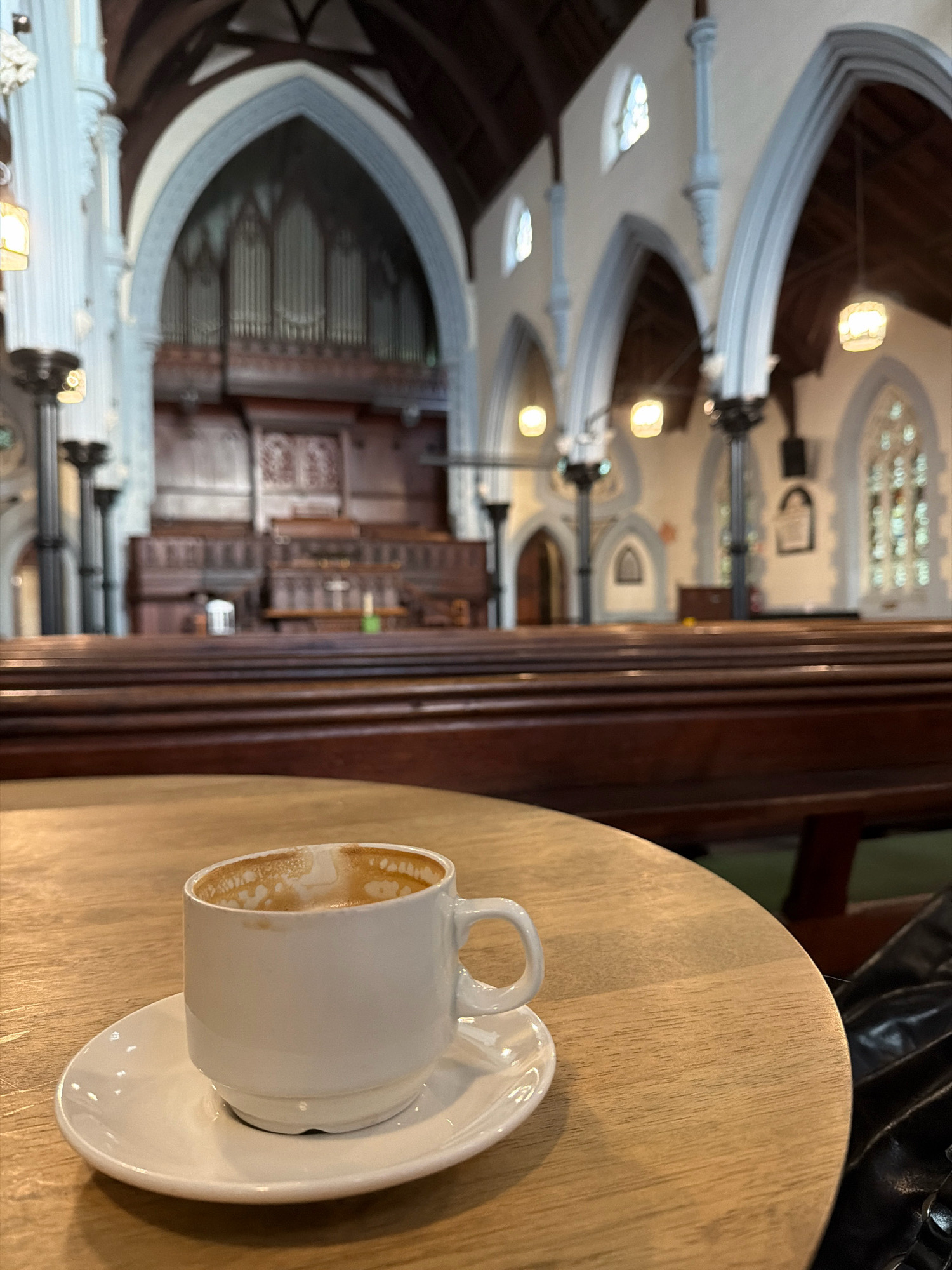 A close-up, eye-level shot focuses on a white ceramic coffee cup and saucer on a light wooden circular table. The cup contains a small amount of coffee residue on its interior. The table is positioned in the foreground, with the background blurred, revealing the interior of a grand, old church. The church architecture features high, arched ceilings with Gothic-style ribbed vaults, large stained-glass windows, and rows of dark wooden pews. In the distance, a pipe organ is visible. The lighting in the church is dim, with warm light emanating from hanging fixtures. The overall impression is one of quiet contemplation within a historic setting. Based on the architectural style and context, this image was likely taken in St. George's Cathedral in Cape Town, South Africa, which is known for its similar interior design.
