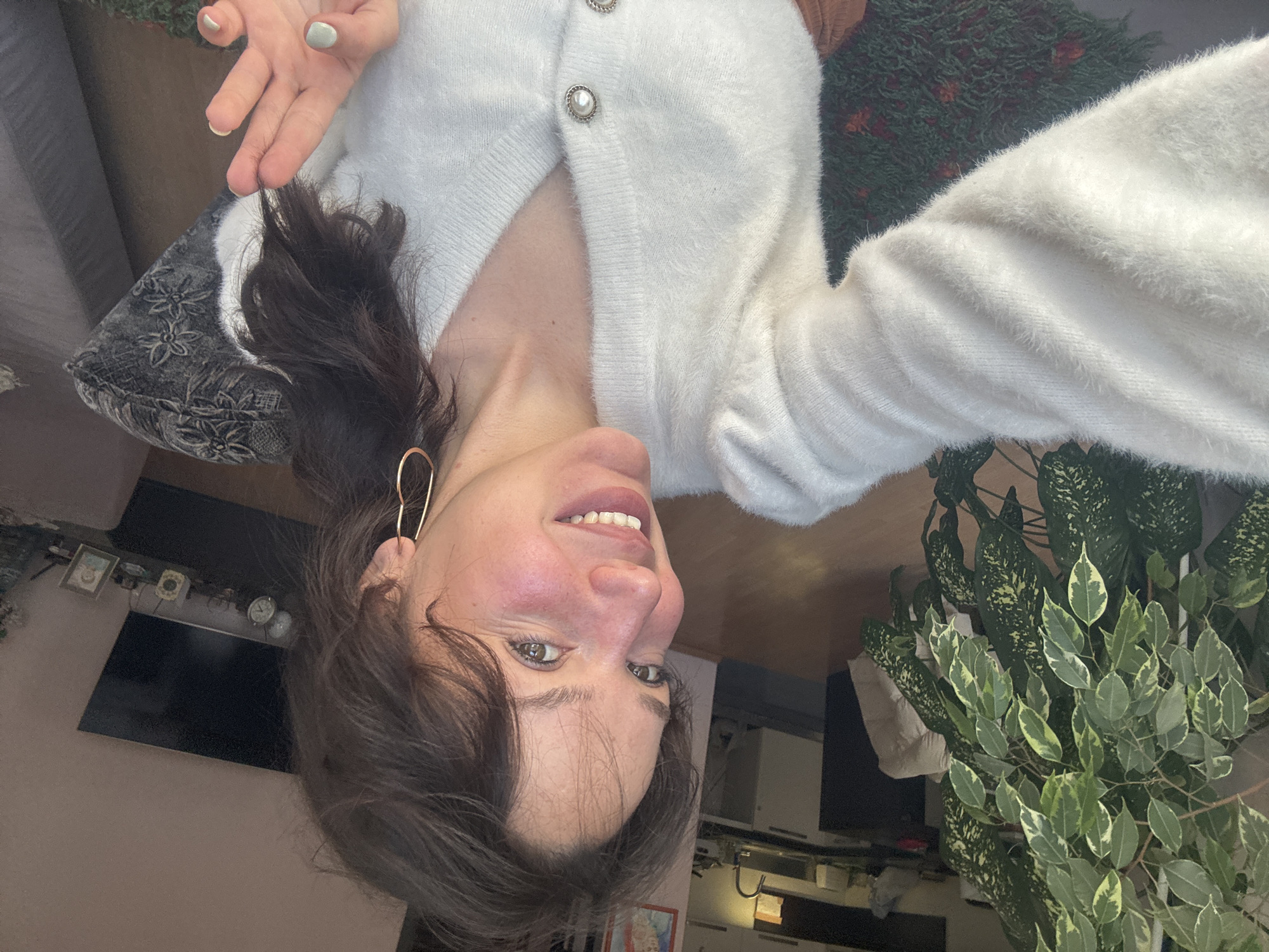 A woman is taking a selfie indoors. She is wearing a white cardigan with pearl buttons. The location appears to be a domestic interior, possibly in Burgas, Bulgaria, given the mention of the city. A television is visible in the background, alongside other domestic furnishings. A plant is also visible. The woman is smiling and gesturing with her hand.