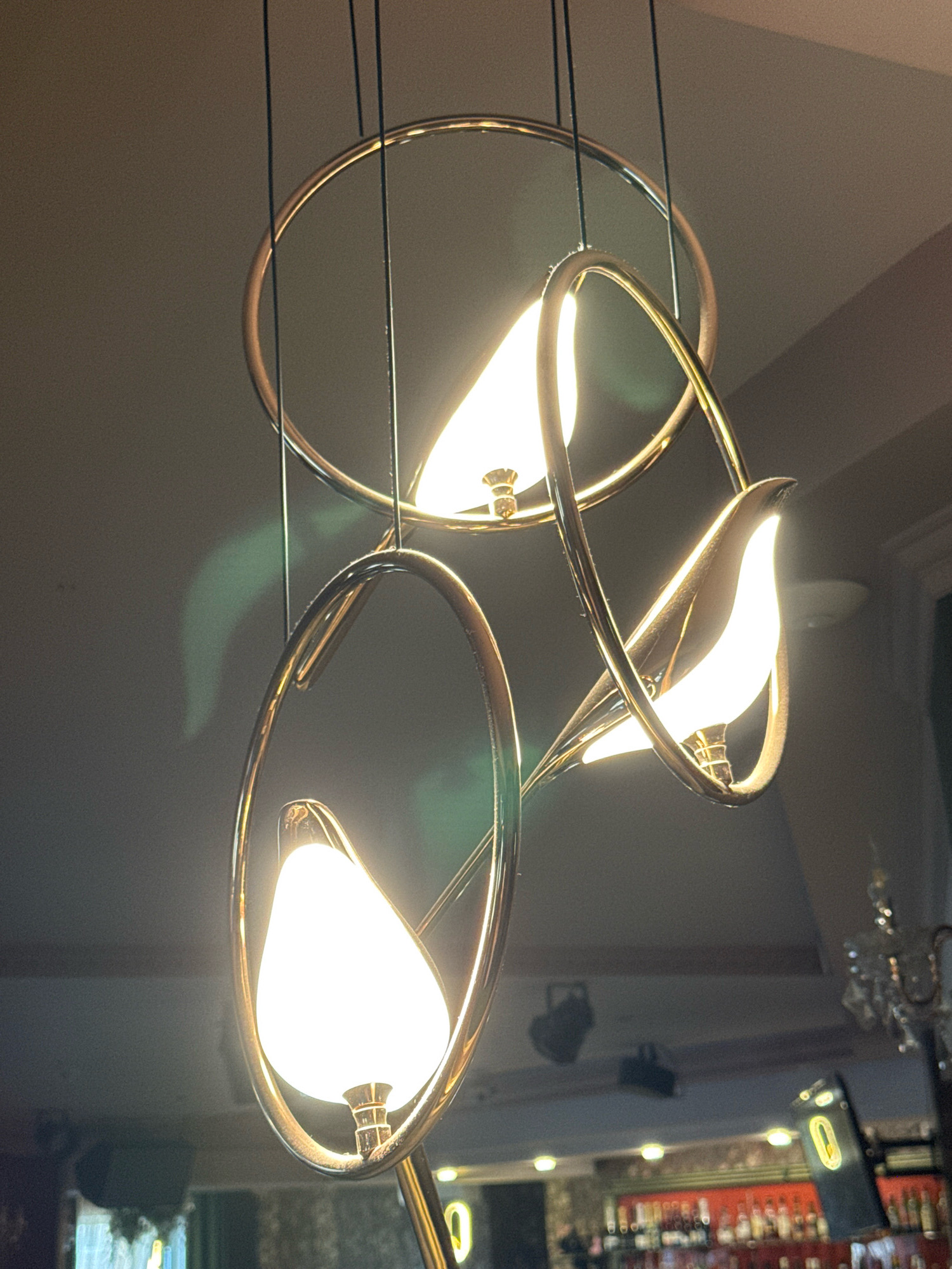 A close-up shot focuses on a modern, gold-colored chandelier hanging from a dark ceiling. The chandelier has a circular frame with three oval-shaped extensions, each containing a frosted white light bulb shaped like a flame. The bulbs are illuminated, casting a bright glow. The background is dark and slightly blurred, with hints of a room below, possibly a restaurant or bar, indicated by the presence of shelves with bottles and lighting fixtures. The scene is captured in Torzhok, Russia.