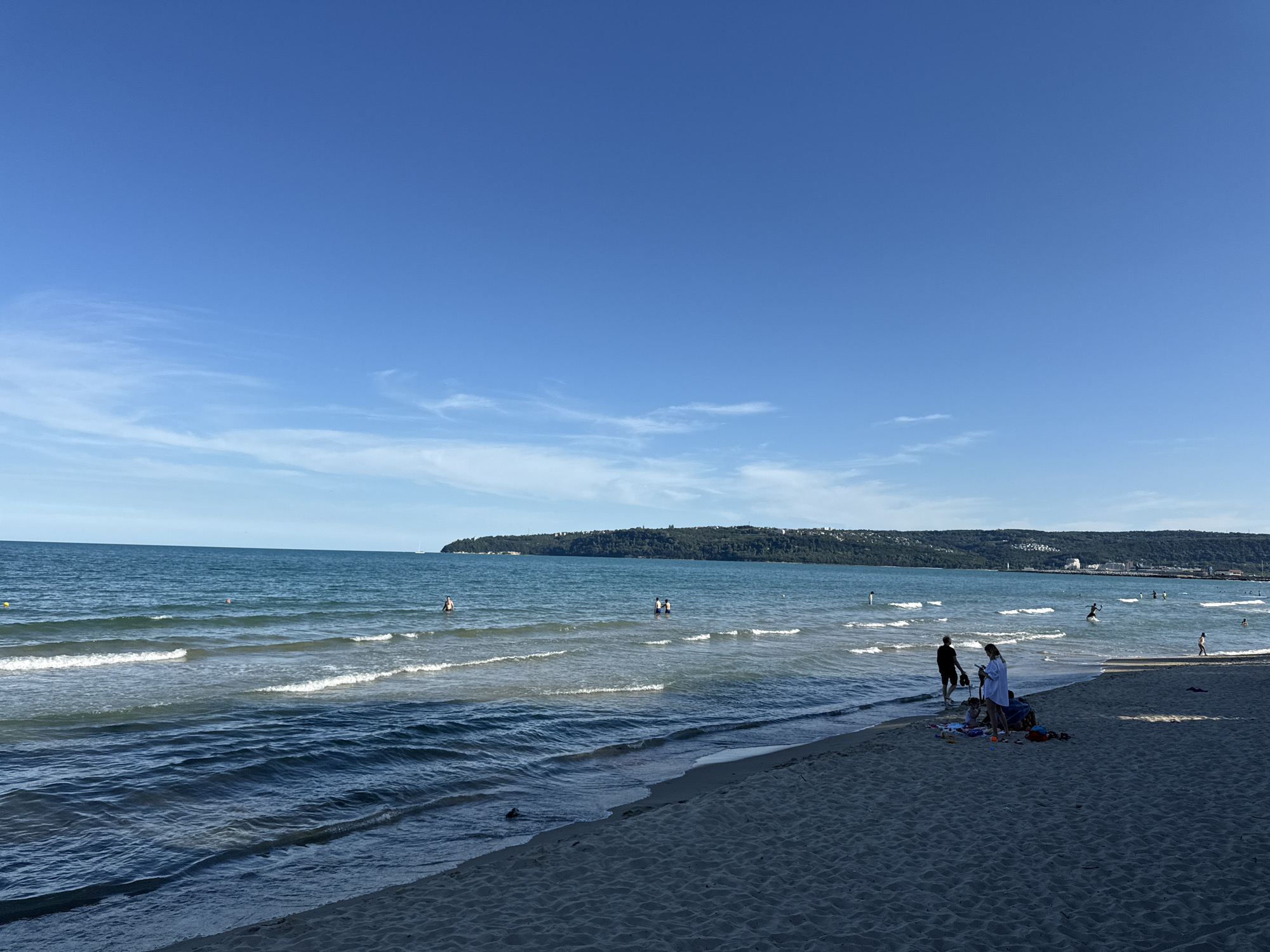 This is a wide shot of the beach in Varna, Bulgaria, on a clear day. The sky is a vibrant blue with some wispy white clouds. The sea is calm and blue with gentle waves rolling onto the sandy shore. There are several people in the water, swimming or wading. On the beach, a few groups of people are relaxing, sitting on towels, or playing. In the distance, a long, tree-covered coastline stretches across the horizon, with some buildings visible on the hills. The overall atmosphere is one of a peaceful summer day at the beach.