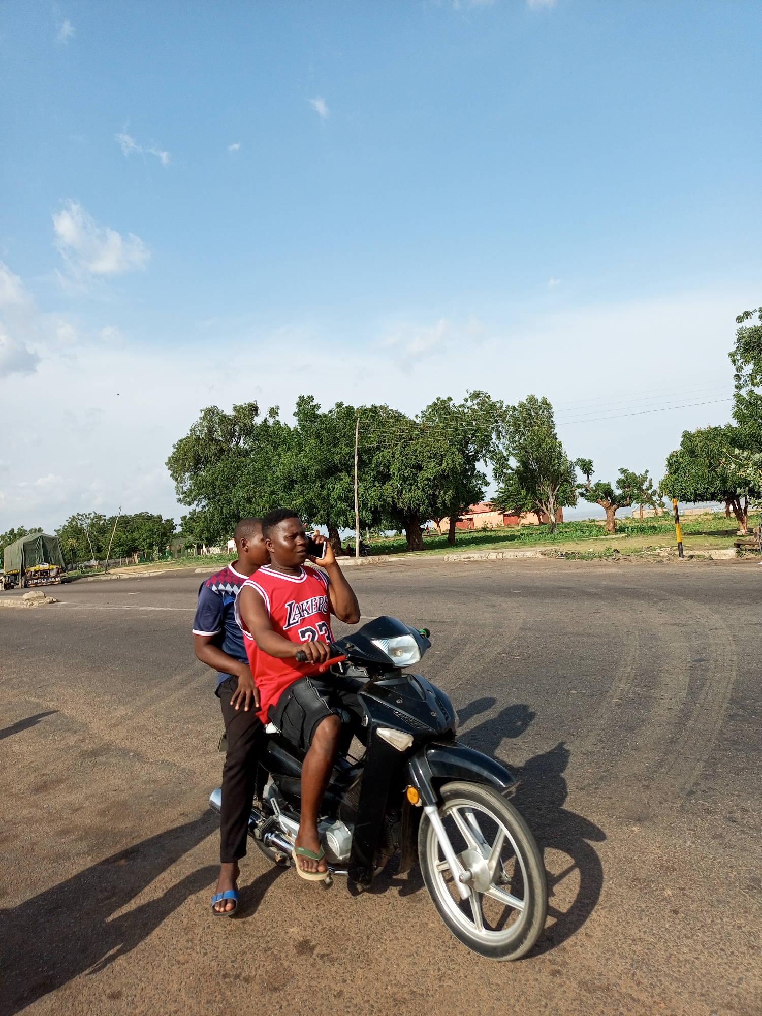 In Gadao, Nigeria, two young men are seated on a black motorcycle. The individual in front, wearing a red Lakers basketball jersey and black shorts, is holding a mobile phone to his right ear, engaged in a conversation. Behind him, another young man, dressed in a blue and white patterned shirt and black pants, is also seated on the motorcycle. The motorcycle is positioned on an asphalt road, casting shadows on the ground. In the background, a truck covered with a green tarp is visible on the left, and lush green trees line the horizon under a bright blue sky with scattered white clouds.