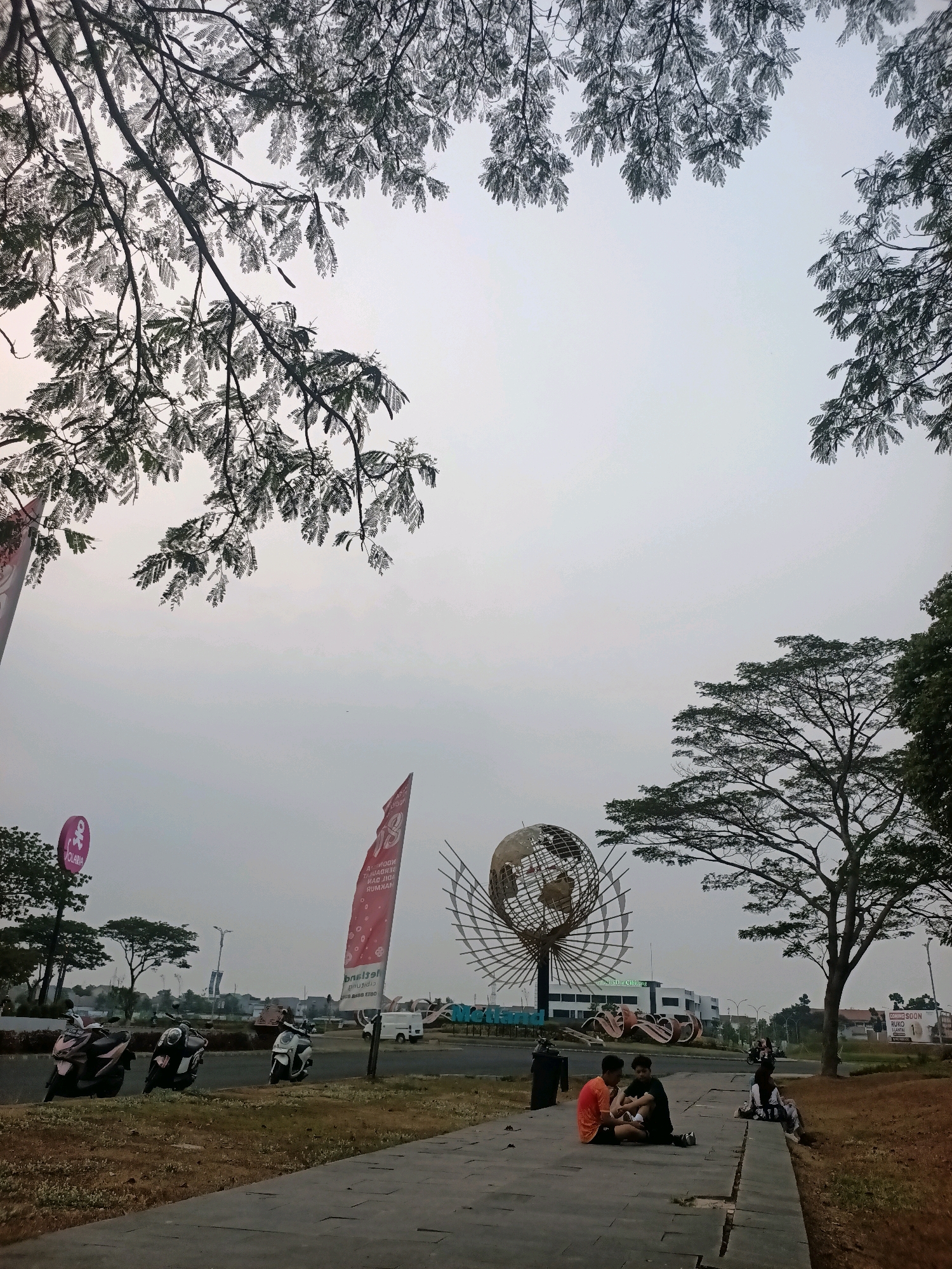 In Telagamurni, Indonesia, a large globe sculpture with an ornate metallic latticework stands prominently in a semi-urban area. The sculpture is positioned on a central island, surrounded by a paved walkway. In the foreground, two young men are seated on the walkway, one wearing an orange shirt and the other a black shirt, appearing to be in conversation. Further to the right, a group of people, including a woman in a white shirt and jeans, are also seated on the walkway, appearing to relax. Several motorcycles are parked along the roadside to the left of the scene. Red flags, one with the word "SOLARIA" and another with text in Indonesian, are visible, along with various modern buildings in the background. Lush green trees frame the sky, with some branches extending into the upper portion of the frame. The sky is overcast, creating a diffused light across the scene.