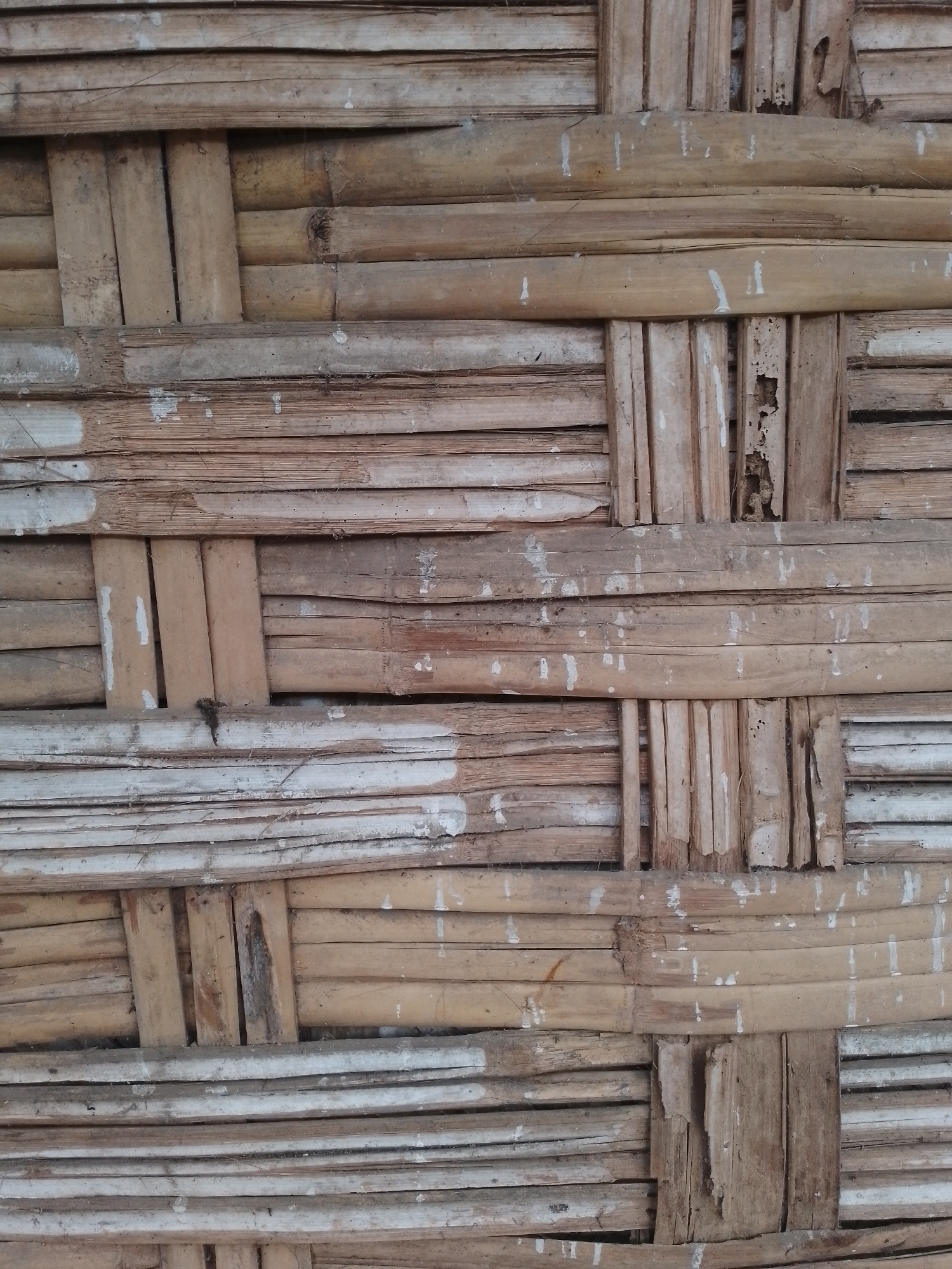 The image displays a close-up, full-frame view of a woven bamboo wall. The bamboo strips are of varying widths and have a weathered, natural color palette ranging from light tan to a more muted grayish-brown. There are visible signs of wear and tear, including some splits and abrasions in the bamboo. Small, irregular splotches of white paint or residue are scattered across the surface, suggesting a previous decorative or functional application. The weaving pattern is a crisscross arrangement, with horizontal and vertical bamboo pieces interlacing to form the structure. The lighting is even, indicating an overcast day or a shaded area, with no harsh shadows. The overall impression is of a rustic and utilitarian building material, commonly found in traditional Indonesian architecture, possibly in a rural or older urban setting such as the Kauman area of Indonesia, known for its traditional crafts and buildings. There are no people, animals, or discernible actions taking place in the image; it focuses solely on the texture and construction of the bamboo wall.