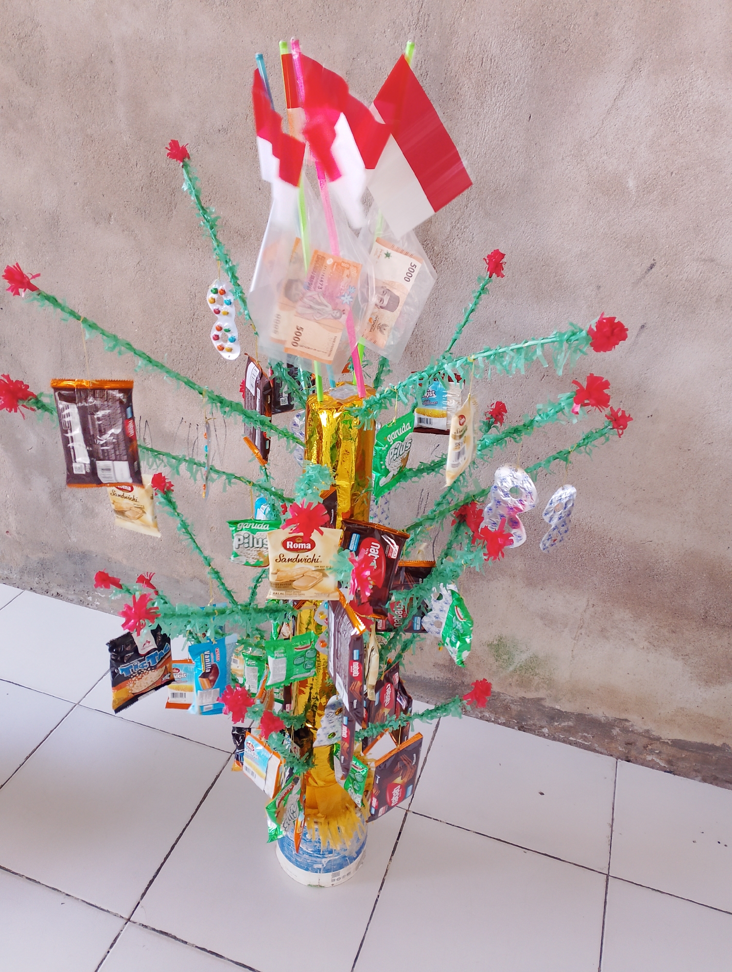 The image depicts a creatively decorated tree, likely for a festive occasion, situated indoors on a tiled floor. The tree's branches are adorned with various items, including Indonesian flags, packaged snacks such as biscuits and chips, and what appear to be paper money, possibly as part of a tradition or game. Red paper flowers are attached to the green branches, adding a decorative touch. The backdrop is a rough, textured wall. The overall scene is brightly lit, and there are no people visible in the frame. The objects suggest a celebration or a cultural event. The location of Bobong, Indonesia, is an urban setting, and the tiled floor and wall are common interior features.
