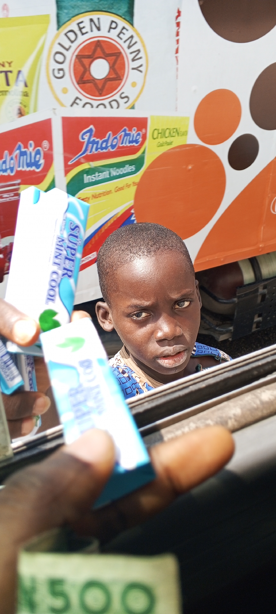 In Agan, Nigeria, a child with dark skin and dark hair looks directly at the camera. The child is wearing a blue and black patterned shirt. In the foreground, a person's hands hold up two small, rectangular blue and white packages of "Super Mint Cool" gum. Below the gum packages, a blurry Nigerian 500 Naira note is visible. In the background, there are signs with product branding, including "Golden Penny Foods" and "Indomie Instant Noodles," along with abstract orange and brown circles. The scene appears to be outdoors, possibly near a food vendor or market stall.