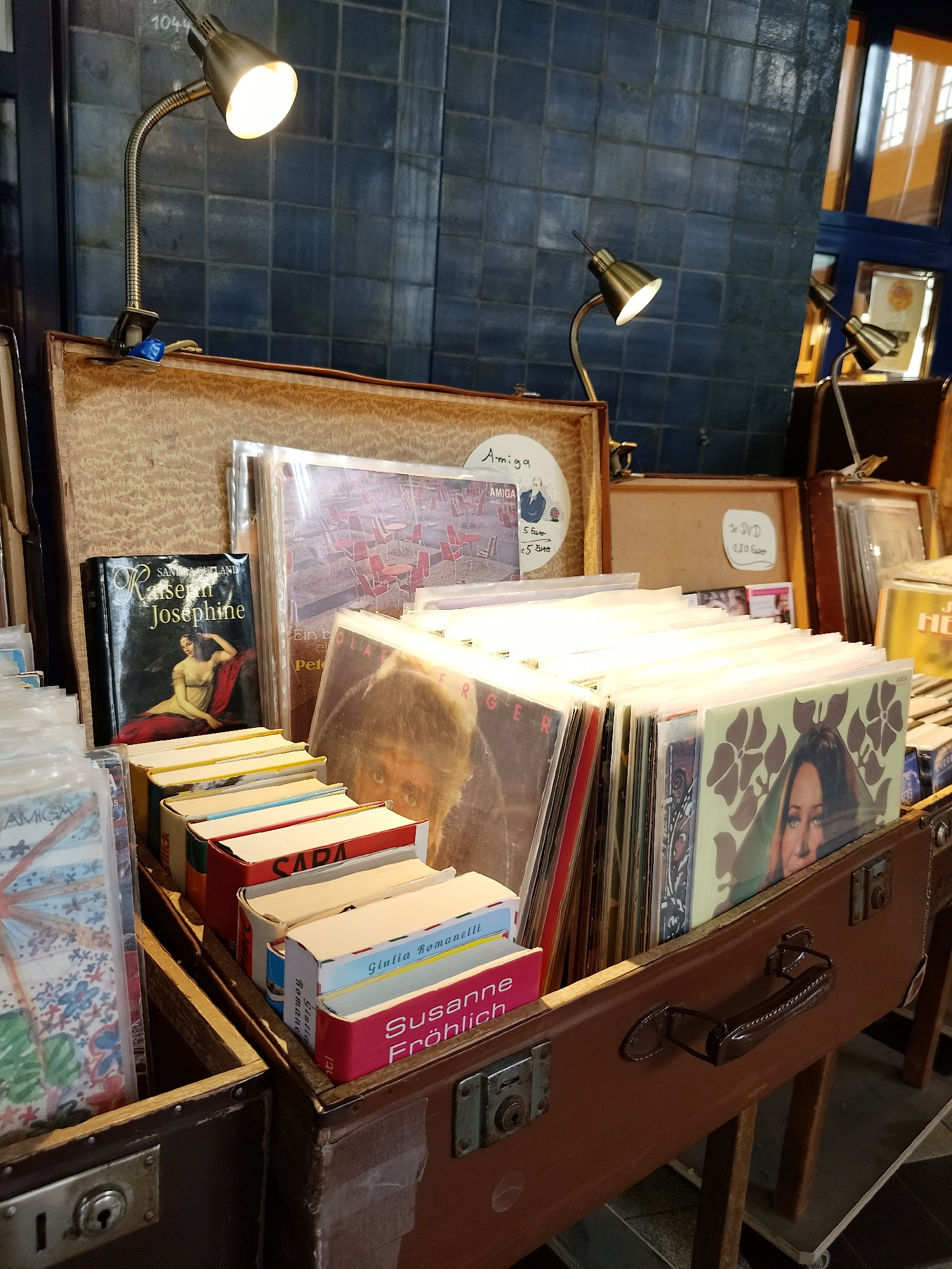 Captured indoors, the photograph presents a street market stall located in Unknown, Unknown. A diverse array of vinyl records and books are neatly arranged in repurposed suitcases, with prices and descriptive labels visible on some items. Titles such as "Kaiserin Josephine," "Giulia Romanelli," and "Susanne Fröhlich" can be discerned. The stall is illuminated by flexible, clip-on lamps that cast a warm glow on the merchandise. The background reveals dark blue, square tiling and a partially visible blue-framed window, contributing to the unique ambiance of the marketplace.