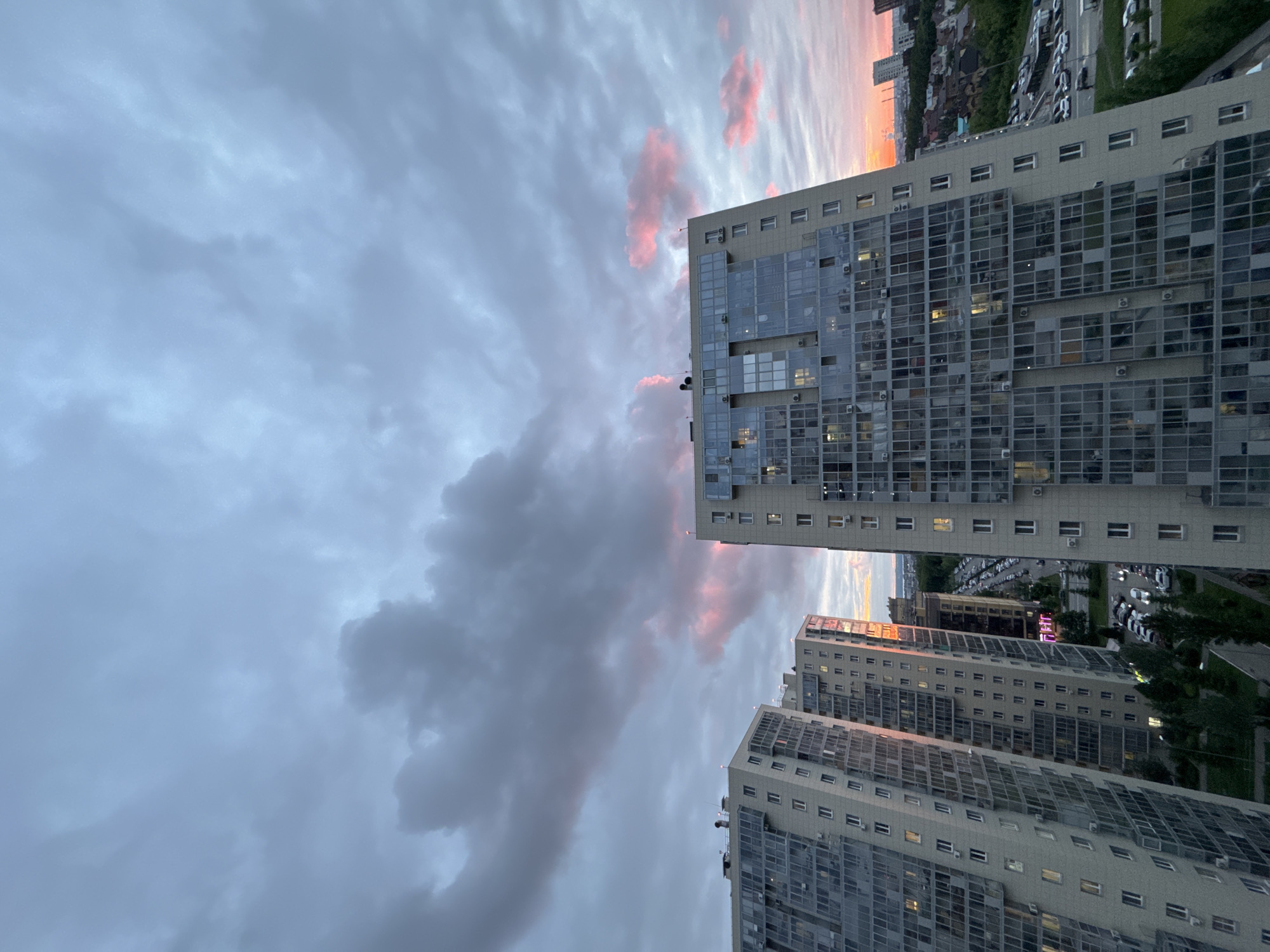 The image shows a view of the city of Kazan, Russia, at dusk or dawn. Tall, modern apartment buildings dominate the right side of the frame, their facades reflecting the soft light. A parking lot filled with vehicles is visible behind the buildings. The sky on the left is filled with dramatic clouds, tinged with pink and orange hues from the setting or rising sun. The image is shot from a high angle, suggesting the photographer was on an upper floor of a building. No people are visible in the shot.