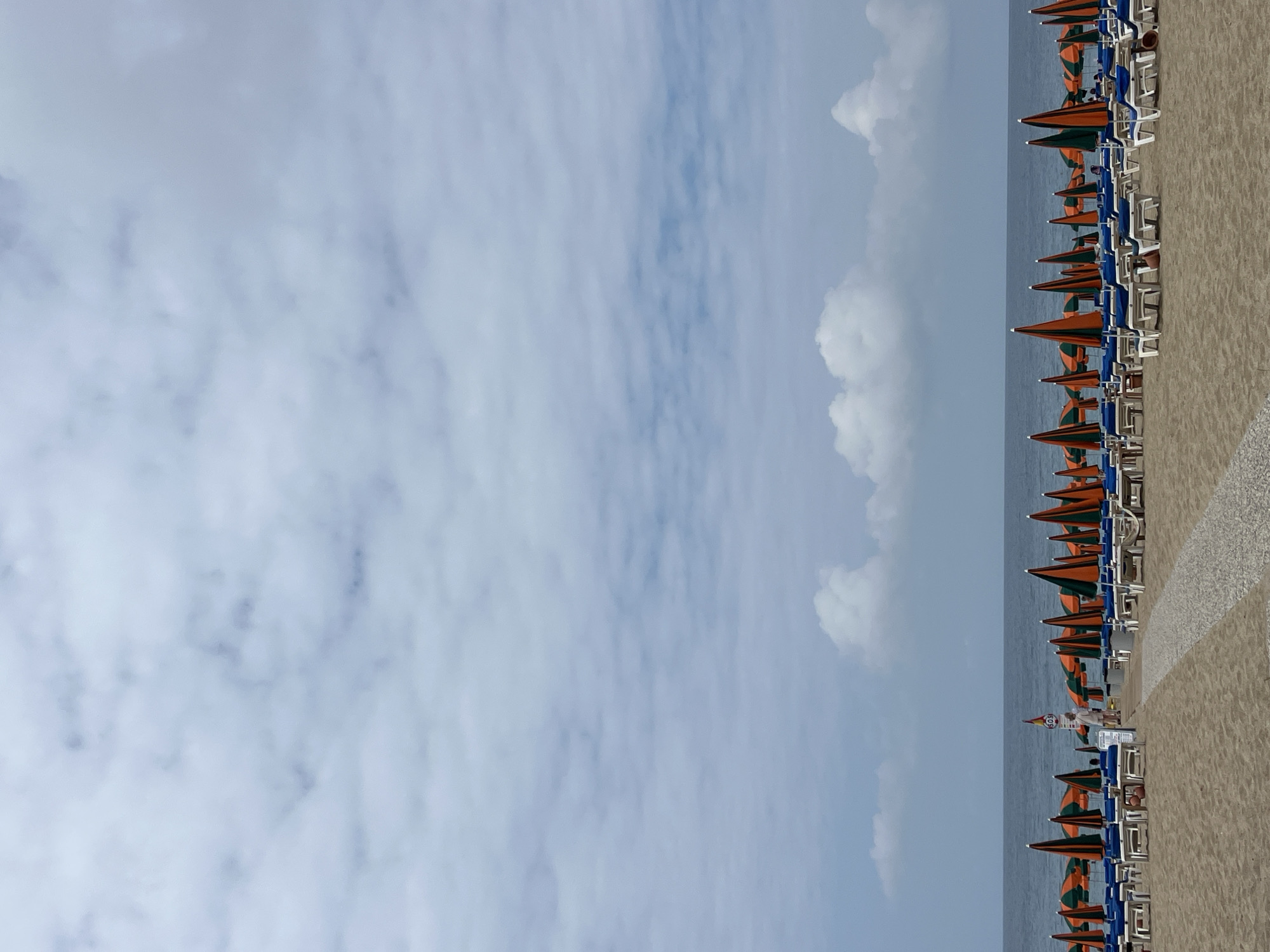 The image shows a beach in Alanya, Turkey. The scene captures a clear sky with scattered clouds, a calm sea, and a sandy beach. A long row of beach umbrellas, alternating in orange and green, extends along the beach, each paired with two white chairs. The beach appears sparsely populated, with no people clearly visible in the scene. The overall setting suggests a peaceful, possibly early morning, beach environment.