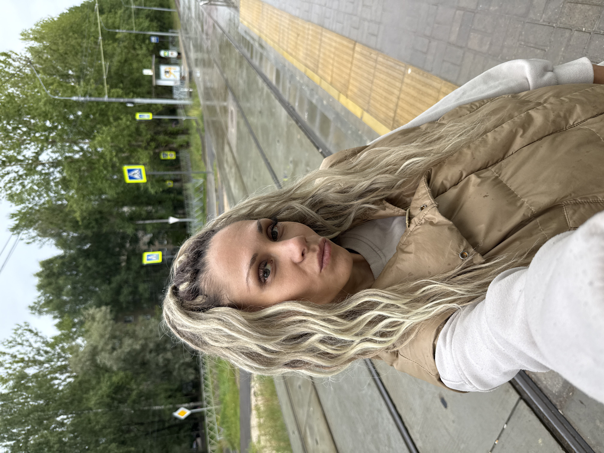 A woman with blonde, wavy hair pulled into a high ponytail takes a selfie outdoors. She is wearing a light-colored sweatshirt and a tan puffer vest. She is standing on a sidewalk next to tram tracks. The ground appears wet, suggesting it may have recently rained. In the background, there are trees, street signs, a bus stop shelter, and overhead wires. The scene is set in the city of Rusanovo, Russia.