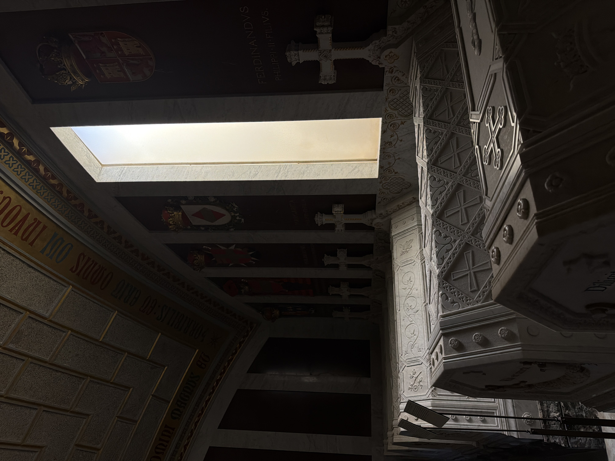 The image depicts the interior of the Royal Pantheon of the Monastery of San Lorenzo de El Escorial, located in San Lorenzo de El Escorial, Spain. The scene features multiple ornate sarcophagi, likely belonging to Spanish royalty, arranged in a dimly lit chamber. The sarcophagi are made of white marble and are elaborately carved with intricate patterns, crosses, and floral motifs. Some of the sarcophagi have inscriptions, though they are largely unreadable in the given lighting conditions. The walls are dark, with some areas featuring marble panels and decorative elements like coats of arms and inscriptions. A prominent feature is a tall, narrow window with light filtering through, illuminating a section of the chamber and casting shadows. Several crosses are positioned on pedestals near the sarcophagi, adding to the solemn atmosphere of the burial site. The ceiling is vaulted and appears to be constructed with a textured material, possibly stone or stucco, with decorative bands. The overall impression is one of grandeur, solemnity, and historical significance, characteristic of a royal mausoleum. There are no people visible in the image.