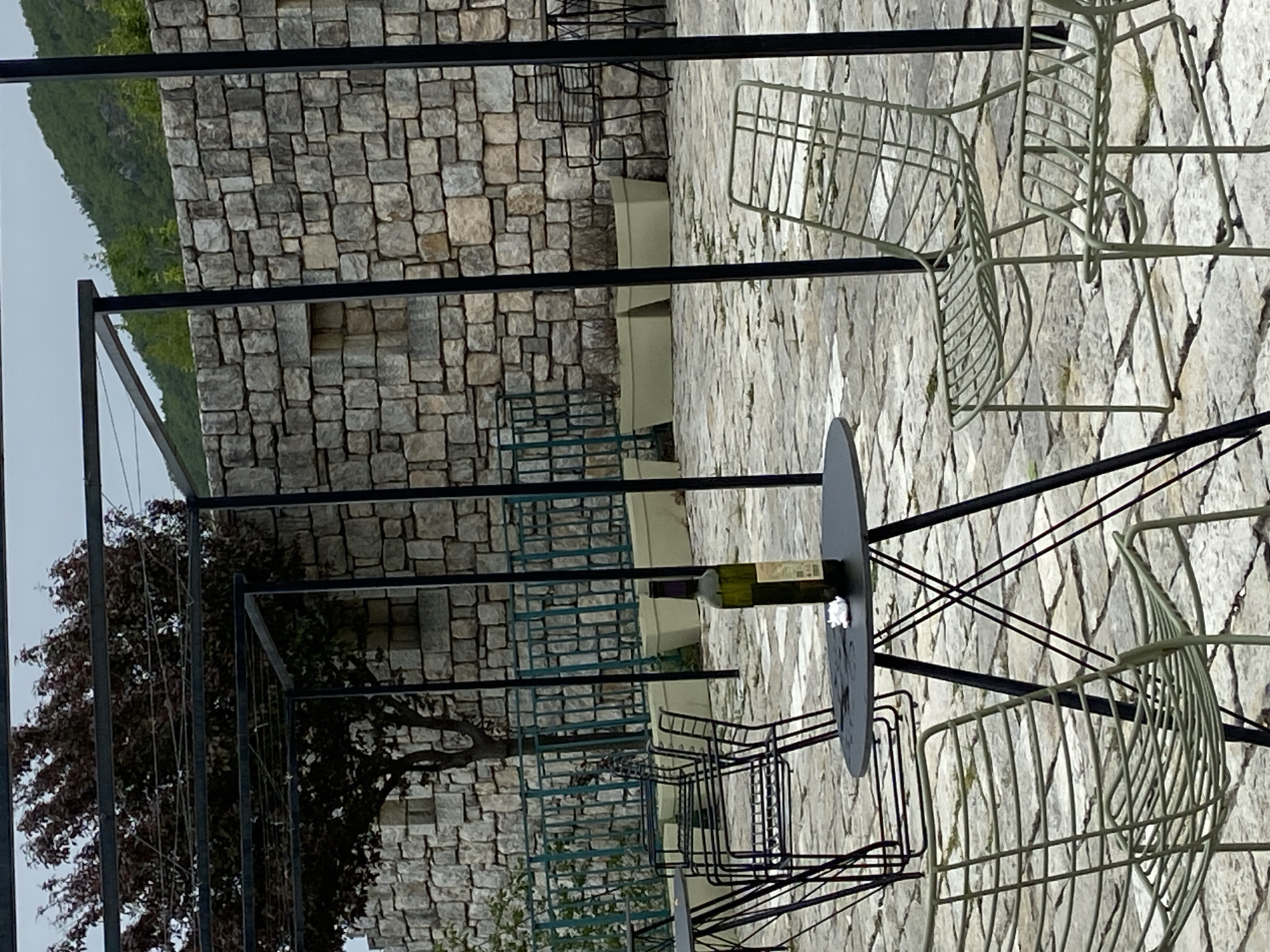 The scene depicts an outdoor seating area in Lipa Dobrska, Montenegro. The background features a stone wall with various chairs and tables. A wine bottle rests on one of the tables. A tree is visible on the left side of the image, and mountains are in the distance. The environment is paved with stone tiles. There are no people visible.