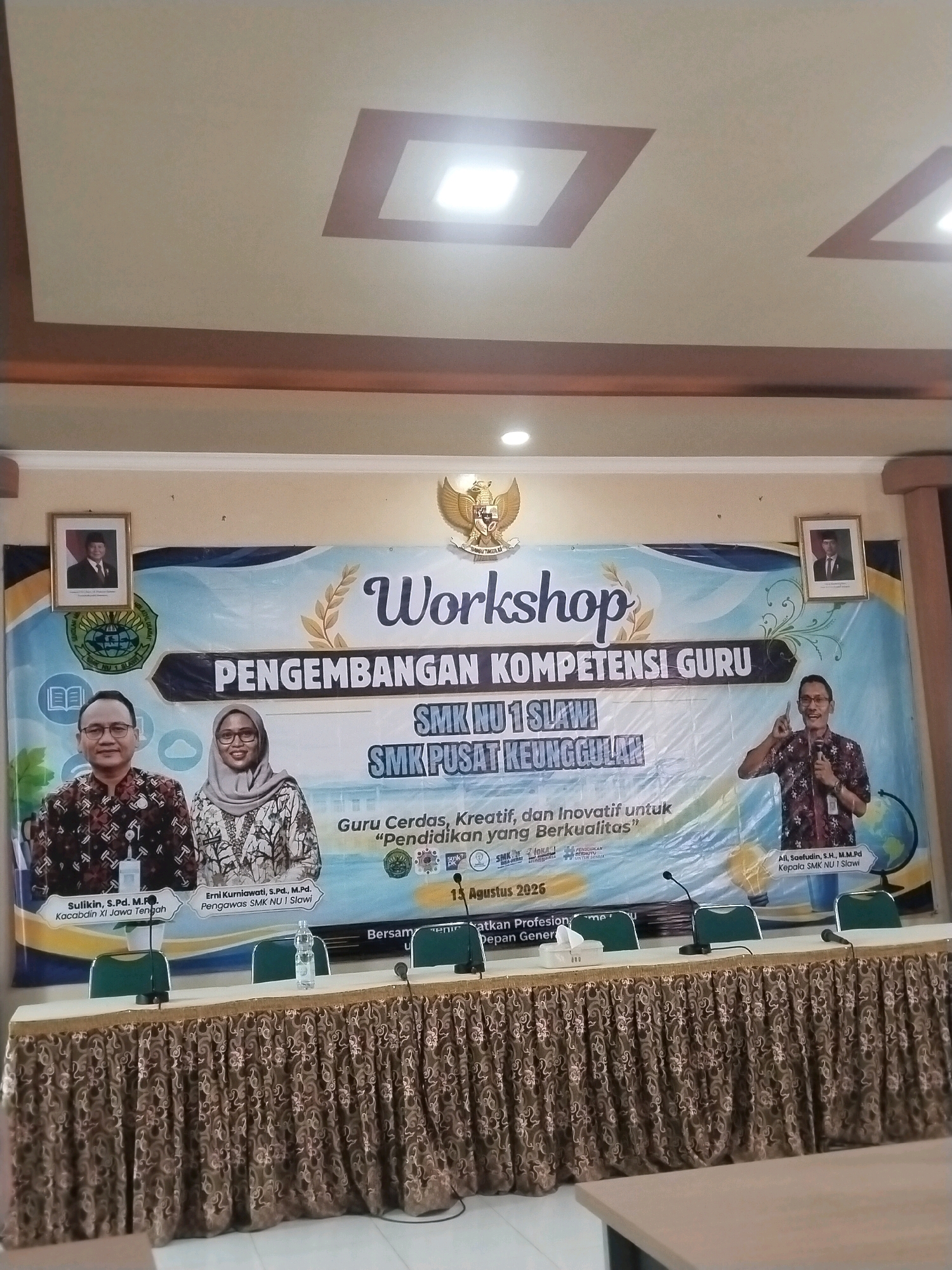 A banner advertising a "Workshop Pengembangan Kompetensi Guru" (Teacher Competency Development Workshop) is displayed on a wall. The banner features text in Indonesian and logos of educational institutions. Three individuals are visible on the banner: a man and a woman on the left, and a man on the right. Two framed portraits of men in formal attire are also on the wall. Below the banner, a table is set up with chairs and microphones, suggesting a presentation or conference setting. The room appears to be indoors, with a ceiling visible in the upper portion of the image. The event is dated "13 Agustus 2026". The location is identified as SMK NU 1 Slawi, with the phrase "Kepala SMK NU 1 Slawi" appearing on the banner, indicating the school's leadership. There is no information in the image to indicate this scene is located in the city of Procot, Indonesia.