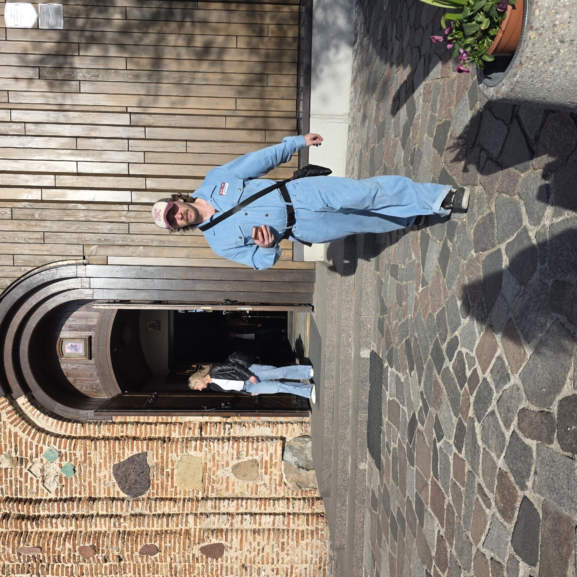 The image, which is rotated 90 degrees clockwise, captures an outdoor scene on a sunny day, likely in a historic urban setting such as Hrodna, Belarus.
In the foreground, a man stands on a paved surface made of irregular stone blocks. He wears a light blue, long-sleeved utility jumpsuit with a black cross-body bag. He has curly dark hair, a mustache, and a light-colored baseball cap. He holds a smartphone in his right hand and looks directly at the camera with a neutral expression. A red and white name tag on his chest reads "HELLO MY NAME IS ANIA" (or ANNA).
Behind the man is a building with a textured wooden facade featuring horizontal planks, positioned above a rustic foundation of red brick and large, interspersed stones. A prominent, large arched wooden doorway with multiple concentric rings serves as the building's entrance, accessible via stone steps.
Inside the doorway, a blonde woman is partially visible, wearing a black leather jacket, a white t-shirt, and light blue jeans. She appears to be looking downwards or to the side.
Strong, distinct shadows fall across the paved ground and the building, indicating bright sunlight, likely during midday or early afternoon. In the upper right corner, a grey planter with green foliage and purple flowers is visible. Two white plaques are mounted on the wooden facade above the man, though their specific text is illegible. The architecture, including the stone paving and the old building materials, aligns with the location context of Hrodna.
