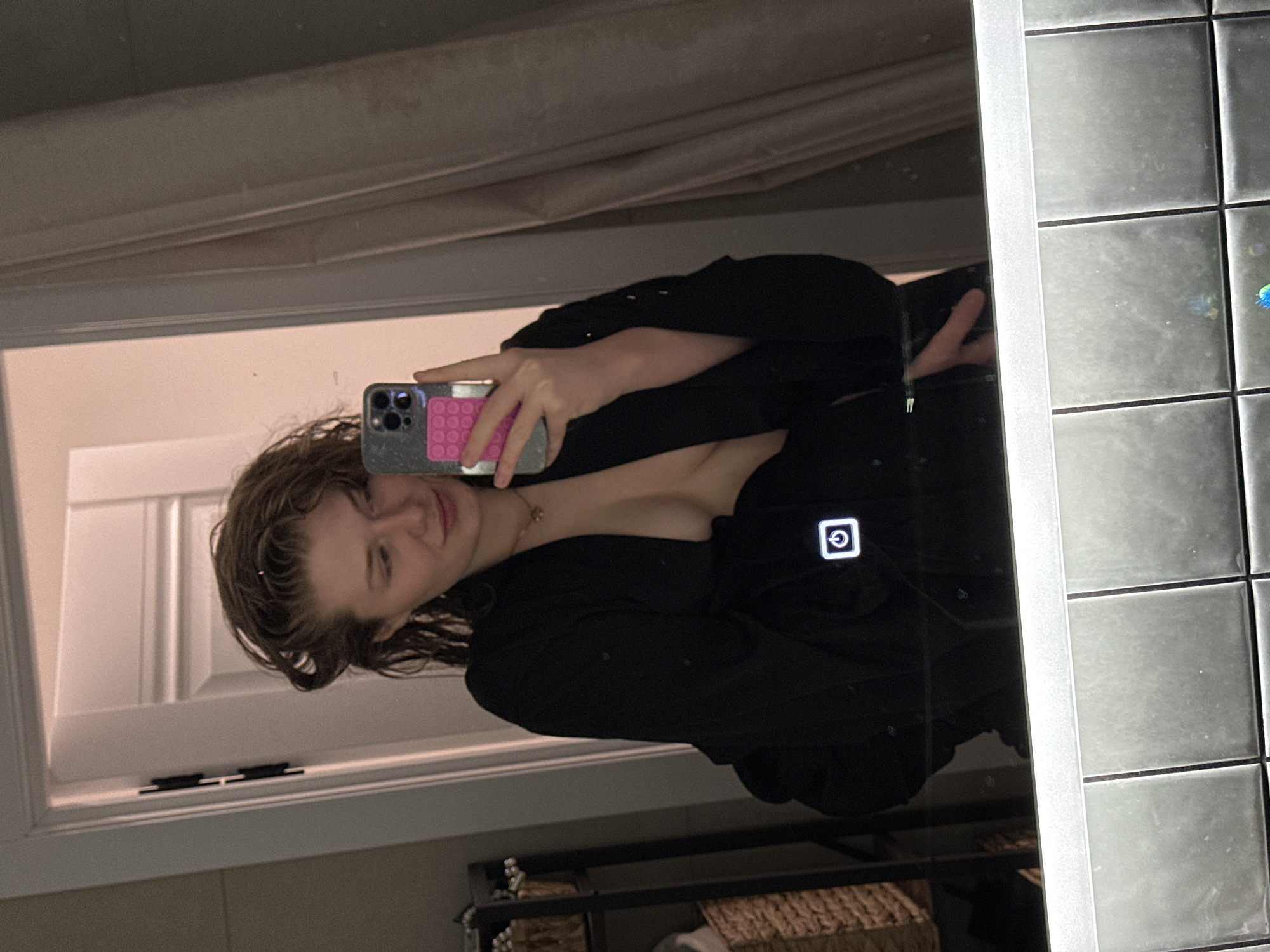 A young person is taking a selfie in a bathroom mirror. The person is holding a smartphone with a pink pop socket on the back. The person has wet hair and is wearing a black garment that exposes some of their shoulder and chest. The bathroom features gray tiled walls and a window with white trim. The environment appears to be dimly lit. There is no indication of this being located in the city of Verhnij Uslon, Russia, or that it even is Russia.