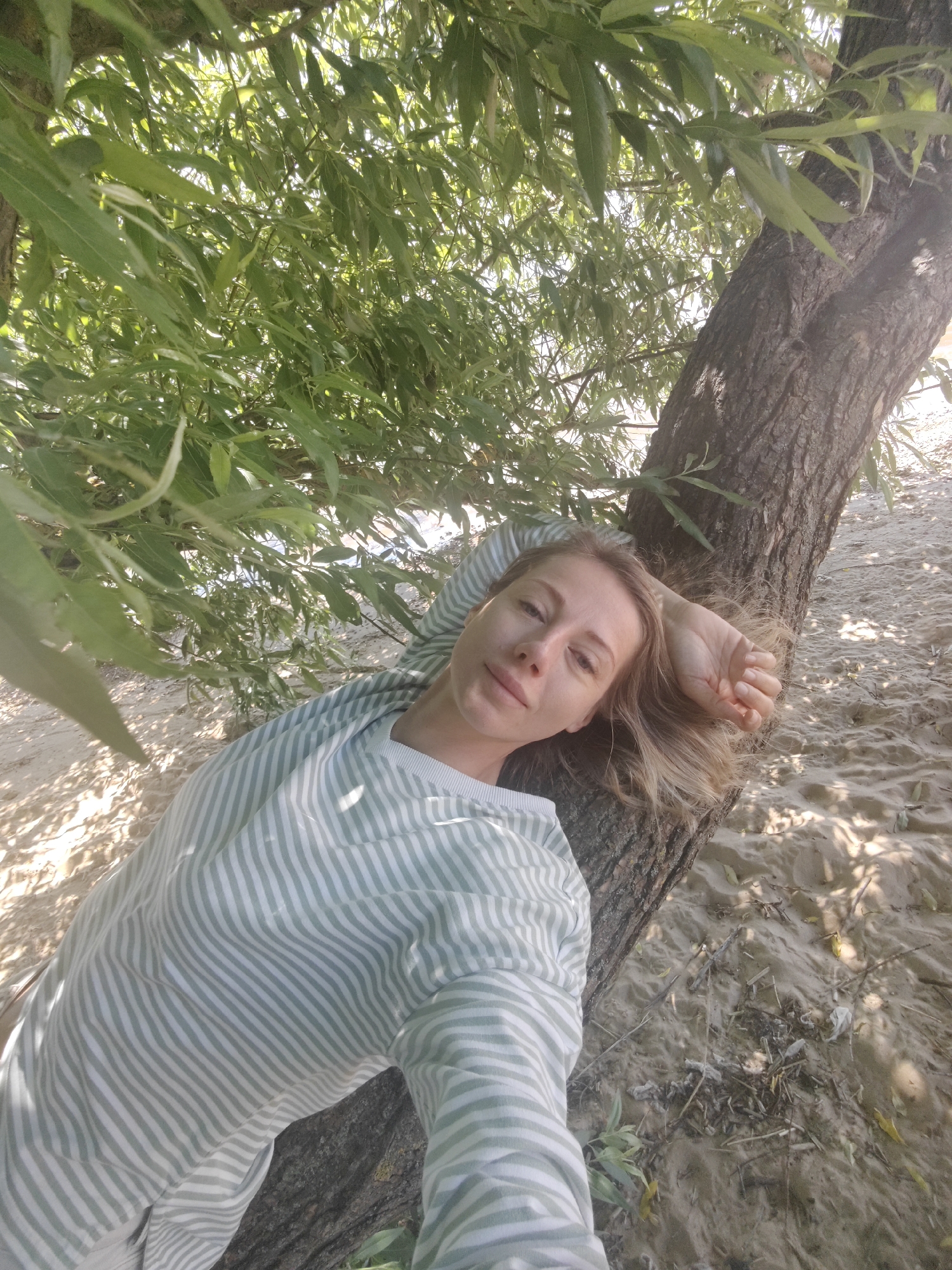 A woman is lying on the sandy bank of a body of water, leaning against a tree. She is wearing a horizontally striped shirt. The leaves of a tree hang overhead, casting dappled shadows on the sand. The image is taken from a high angle, looking down at the woman. The location is not Saint Petersburg, Russia.