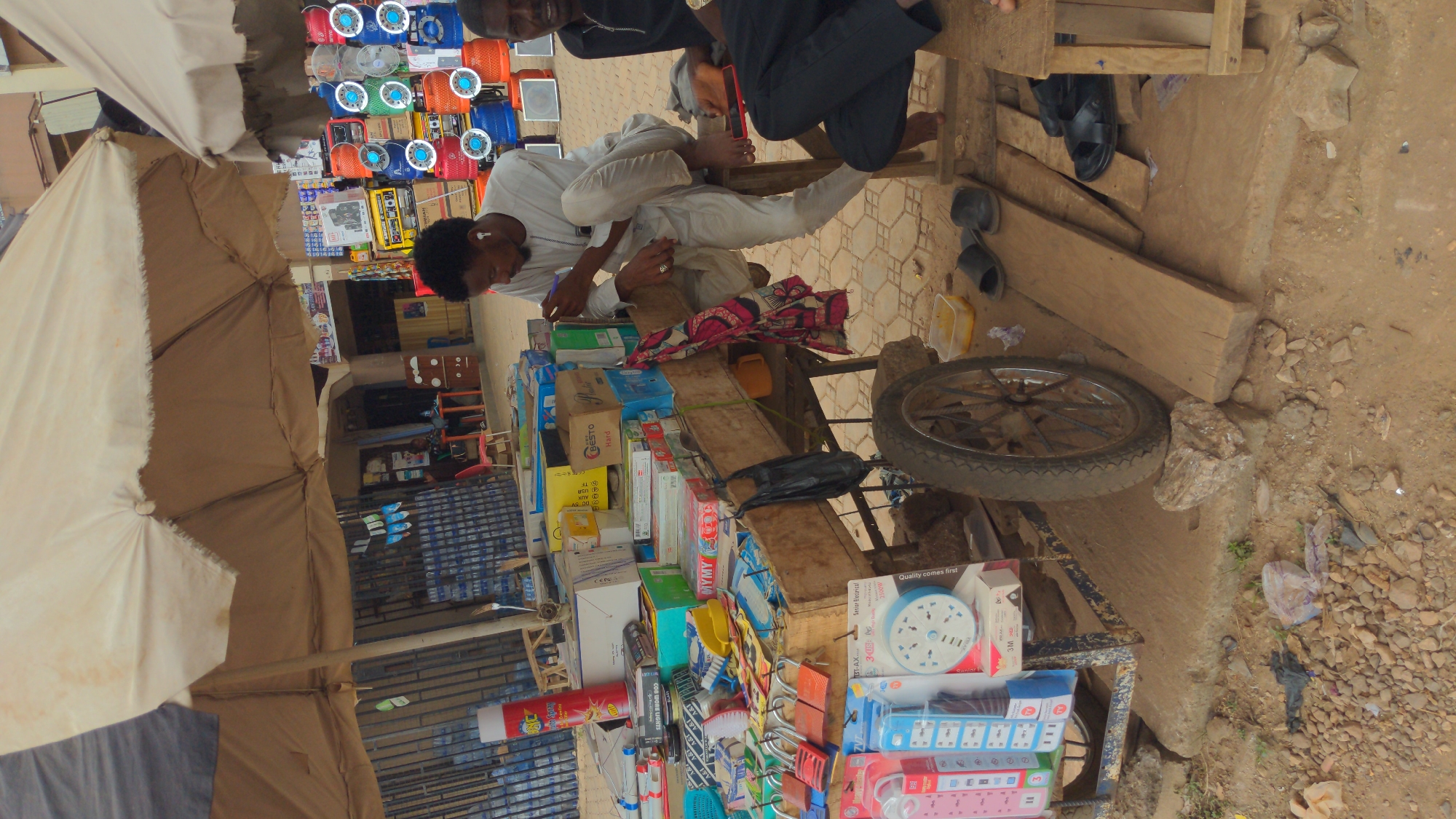 A street scene in Ado, Nigeria, shows two men sitting on wooden planks behind a cart laden with various household and electrical items. The man on the left wears white and appears to be writing on a notepad, while the other man wears black. A large umbrella shades the cart, which displays items like extension cords, lightbulbs, clocks, and boxes of various products. Background shows various shops and electronics supplies. The ground is dirt with trash.