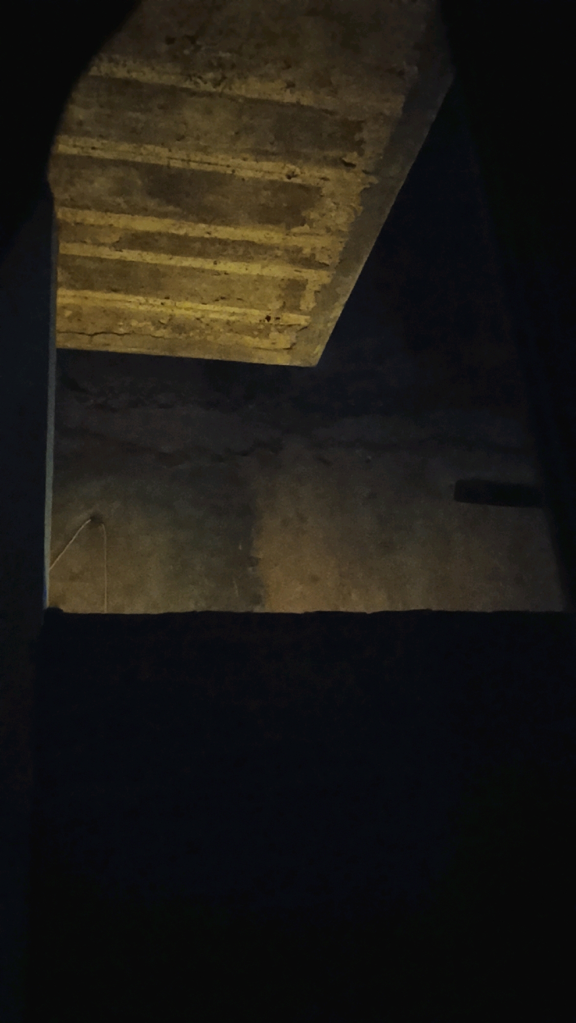 The image shows a concrete structure, possibly a ceiling or overhang, with visible rebar or support beams. The surface is rough and weathered, with some yellow discoloration. Below the concrete structure, there is a wall or surface that appears to be made of rough-textured concrete or stone. A thin wire hangs down from the upper structure towards the wall. The scene is dimly lit, with shadows obscuring much of the detail. The overall impression is one of a dark, possibly underground or enclosed space. The location is identified as Ujungrusi, Indonesia, but no specific details about the site are provided.