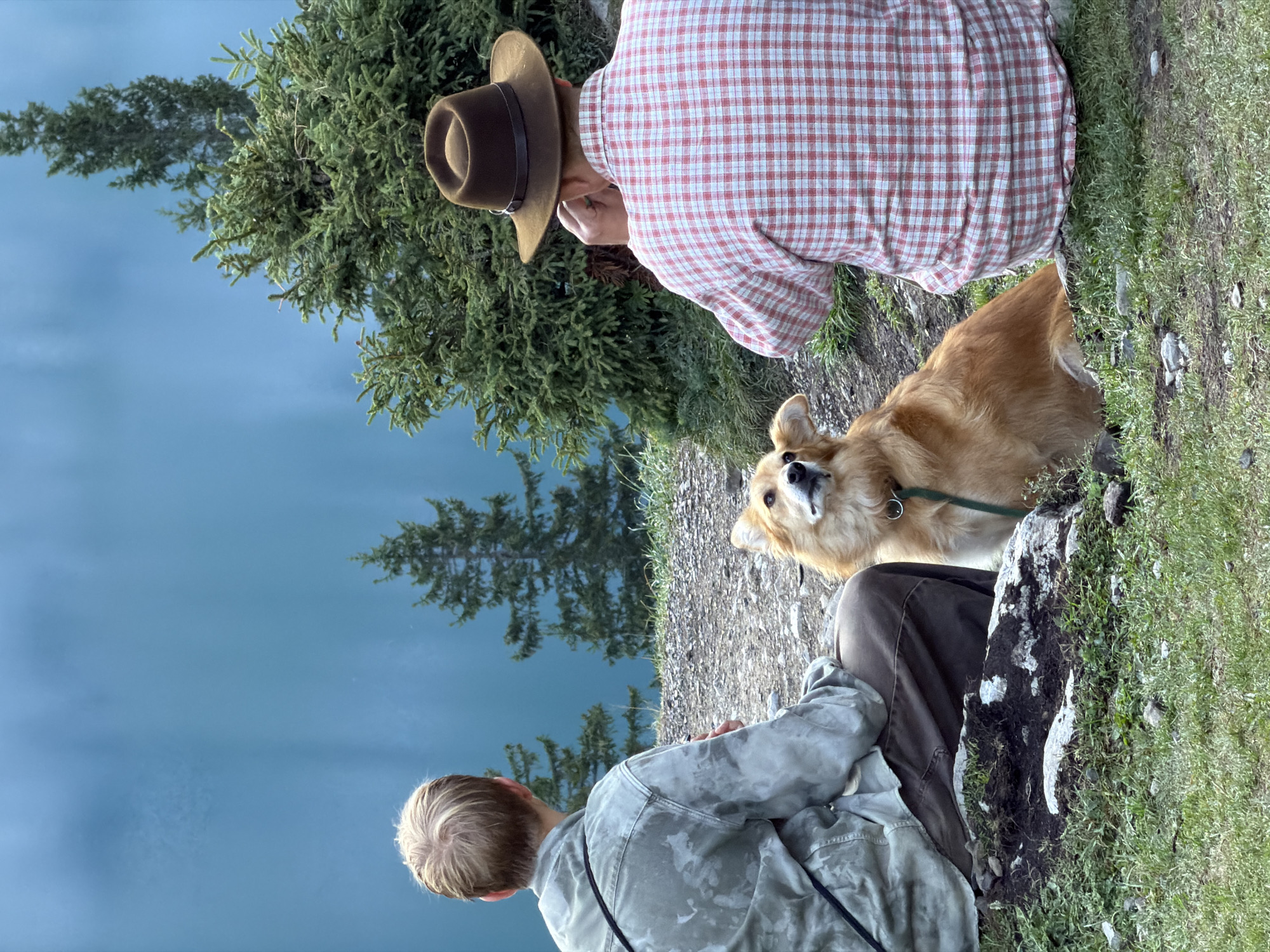 Two individuals, along with a dog, are seated on a rocky, grassy incline overlooking a large body of calm, blue water. The person on the left, viewed from the back, has short blond hair and is wearing a light-colored, camouflage-patterned jacket over dark pants. A black strap crosses their left shoulder. The dog, a medium-sized golden-brown breed, is seated in front of and between the two individuals, looking upwards and to the left. It wears a green collar and leash. The person on the right, also viewed from the back, is wearing a red and white checkered shirt and a brown hat with a dark band. This individual appears to be raising their right hand to their face, possibly adjusting their hat or sunglasses. The background consists of dark green evergreen trees and the expansive blue water. The scene suggests a scenic overlook, likely in a natural environment outside of the immediate city center of Kandersteg, Switzerland, given the expansive natural landscape.