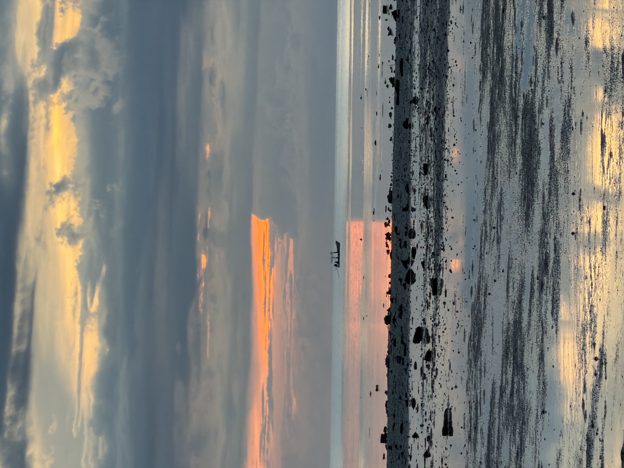 A wide shot captures a serene coastal landscape at dusk in Baan Plai Laem, Thailand. The sky is a dramatic blend of dark grey clouds and vibrant streaks of orange and yellow, hinting at the setting sun. A small, silhouetted fishing boat is visible in the middle distance on the calm ocean. The foreground is a rocky, wet shoreline, reflecting the colors of the sky. The scene is quiet and still, evoking a sense of peaceful solitude.