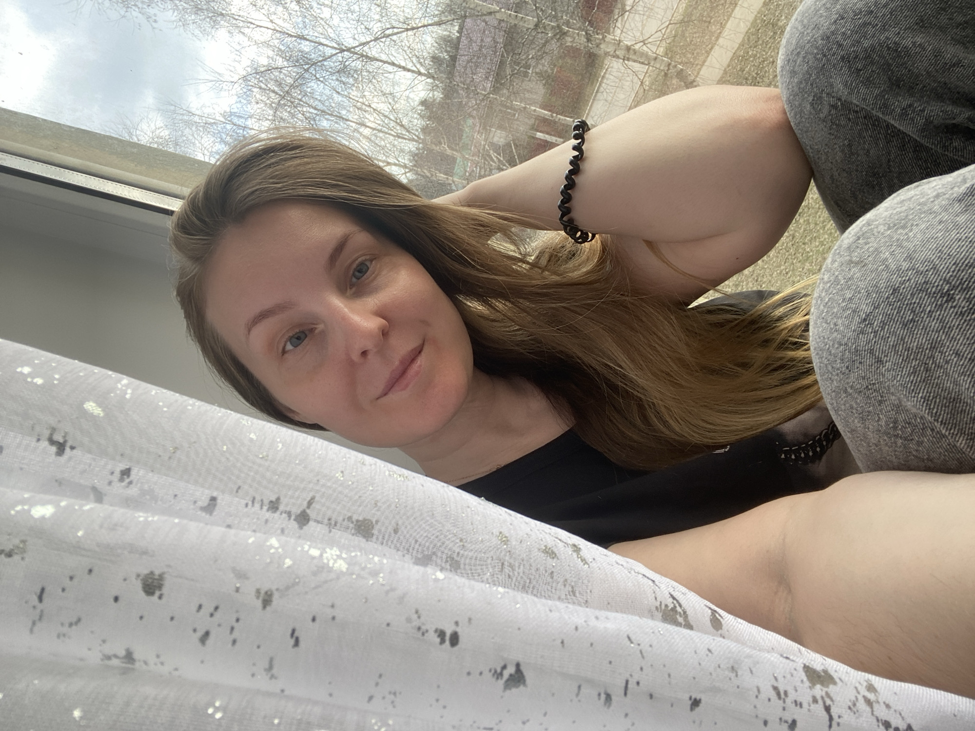 The image captures a selfie of a woman indoors, looking out through a window. The woman has medium-long, light brown hair, fair skin, and light-colored eyes, and she is looking directly at the camera with a gentle, pleasant smile. She is wearing a black top and what appear to be grey denim pants. A black coiled bracelet is visible on her right wrist, as her arm is bent with her hand near her head.
The setting shows a large window providing a view of bare trees and a cloudy sky, suggesting a season like late winter or early spring. Natural daylight illuminates the scene. A white, sheer curtain adorned with silver-grey speckled patterns is visible in the foreground, partially obscuring the bottom left of the frame. The overall atmosphere is relaxed. No visible text is present in the image. The location context provided is Noviy, Russia.