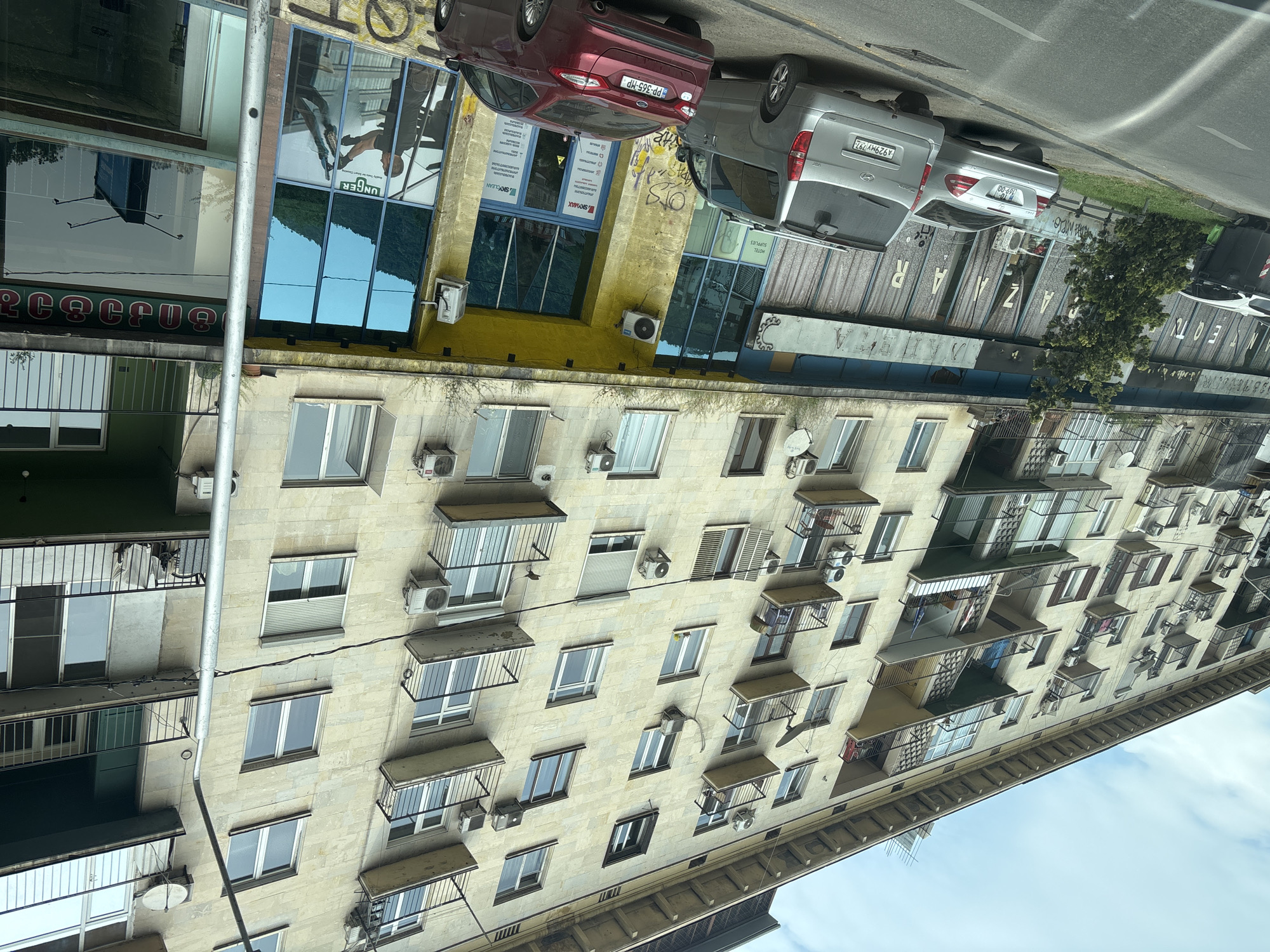 The image shows an apartment building in Tbilisi, Georgia. The building is several stories tall and has many windows and balconies. There are several air conditioning units attached to the building. The building is beige and has a rough texture. A few cars are parked on the street. Some of the businesses at the base of the building are "SKYMAX", "SKYCLEAN", "HOTEL SUPPLIES" and "UNGER."