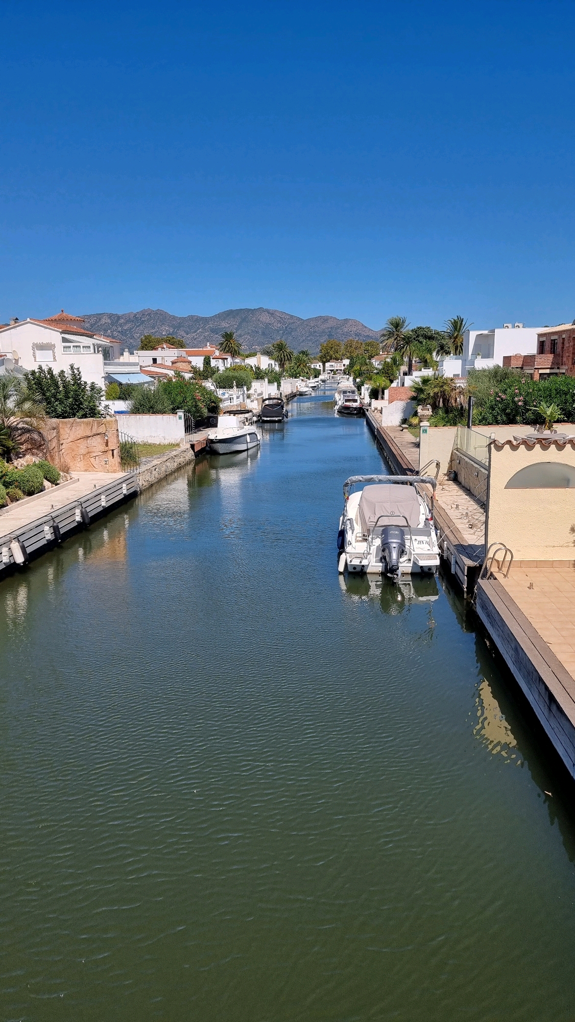 The image captures a sun-drenched canal in the Empuriabrava residential area of Castelló d'Empúries, Spain. The canal, a prominent feature of this coastal town, is lined with modern white villas and lush vegetation, including palm trees. Several boats are moored along the canal's edges and floating on its calm, dark green waters. The sky is a clear, vibrant blue, and distant mountains form a hazy backdrop. The scene is tranquil, evoking a sense of vacation or a leisurely lifestyle, with no people visible.